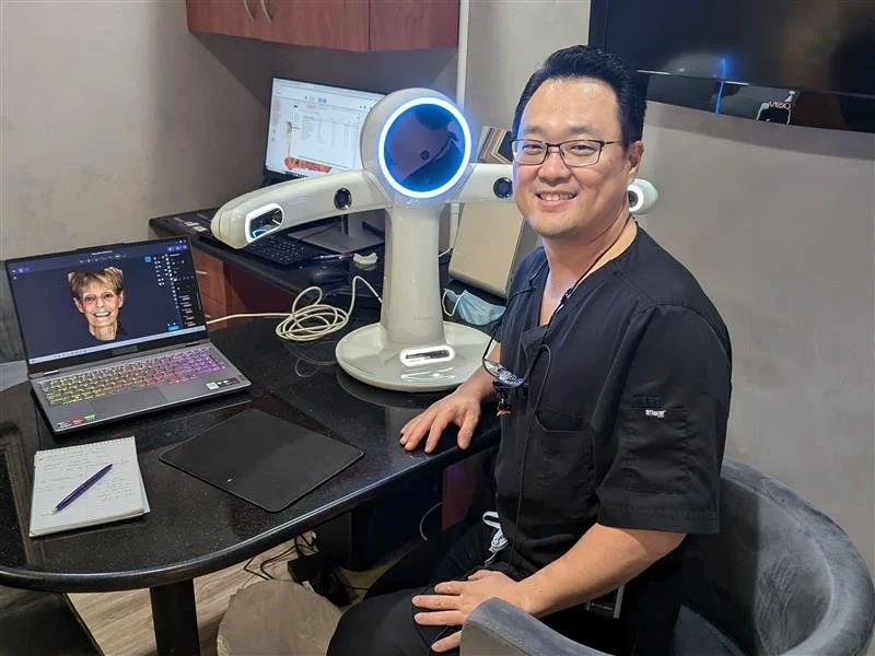 A man sitting at a desk with a smile, wearing black scrubs, in a medical or office setting with medical equipment and a computer, alongside a robot with a circular blue-lit face.