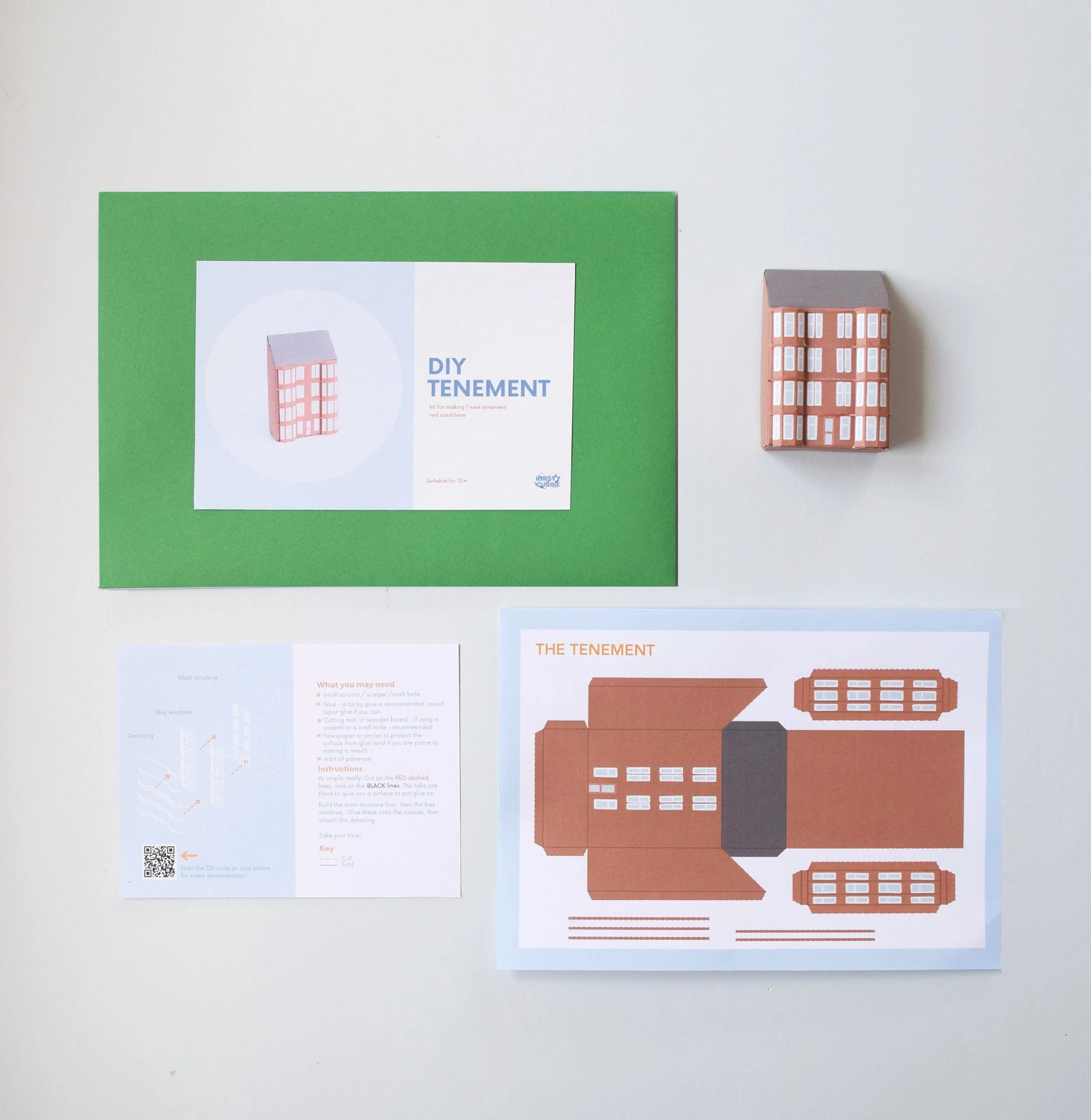 Photograph of various elements laid out flat on a blue background. Includes a green envelope, an instruction sheet, a miniature paper model of a tenement and a sheet with the tenement laid out flat.