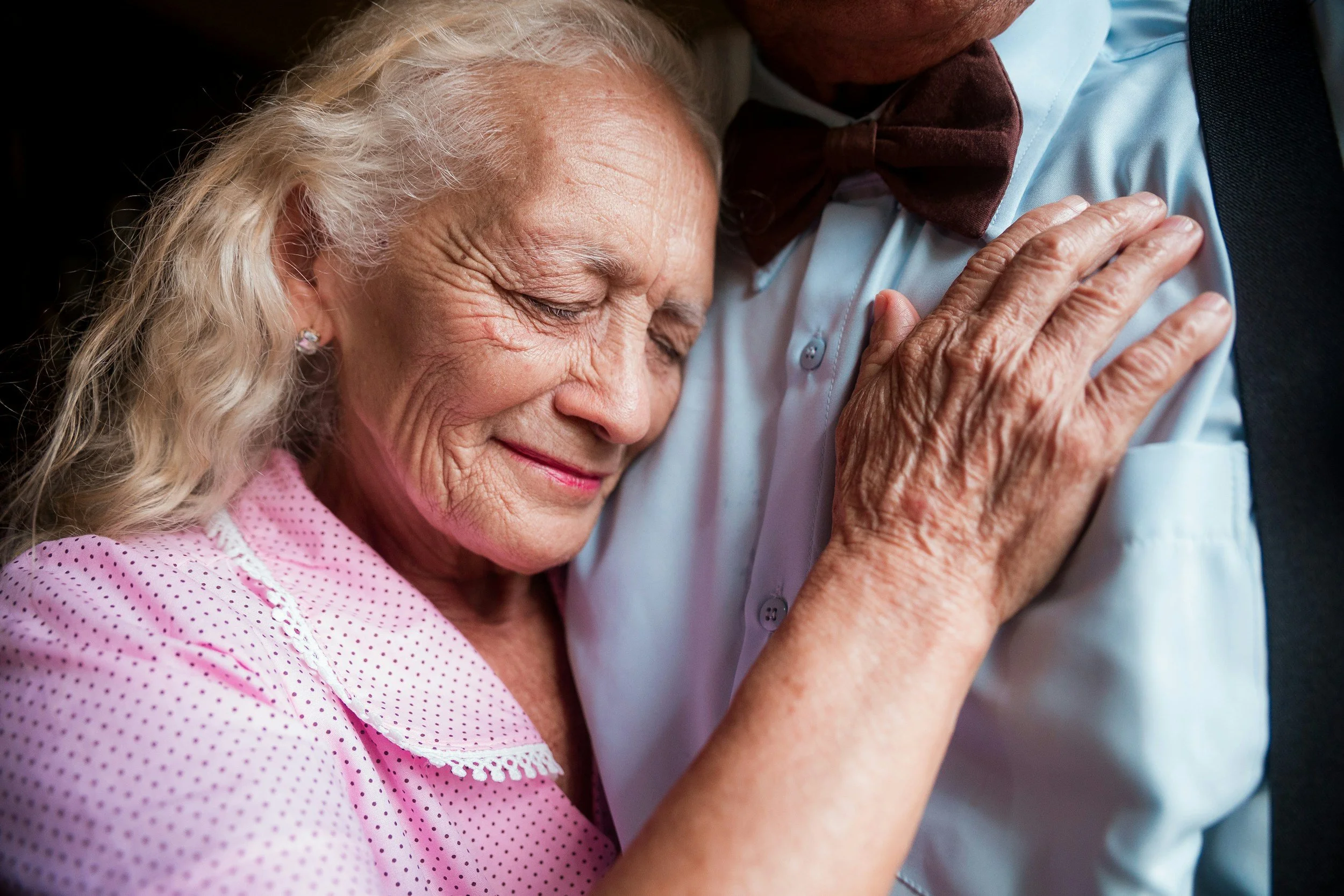 An elderly woman with gray hair and earrings, wearing a pink polka-dot blouse, is hugging a person with a dark suit and brown bow tie, their faces close and eyes closed in a tender embrace.