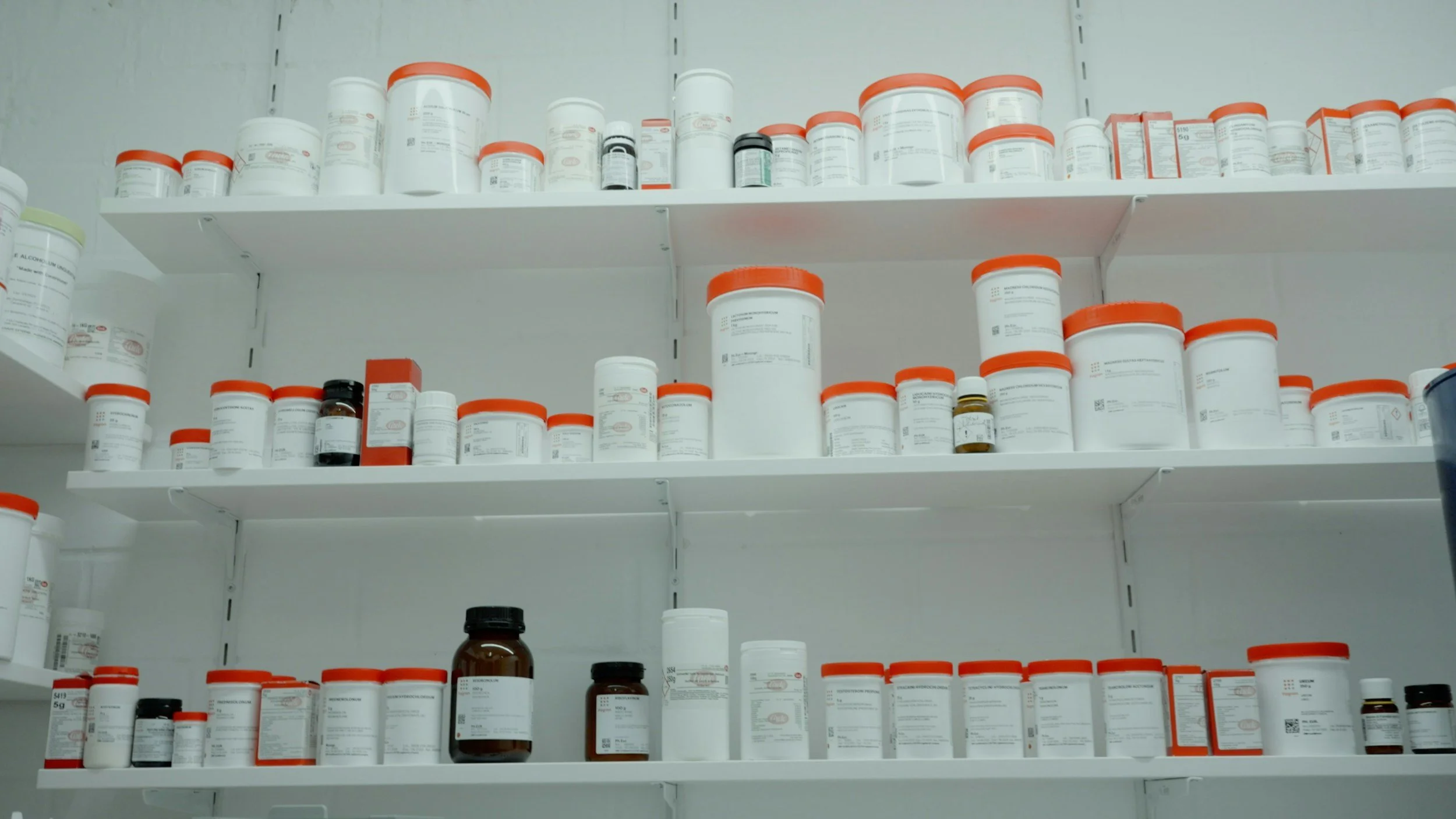 Multiple shelves filled with white prescription pill bottles, some with orange or black caps, in a pharmacy or medical storage area.