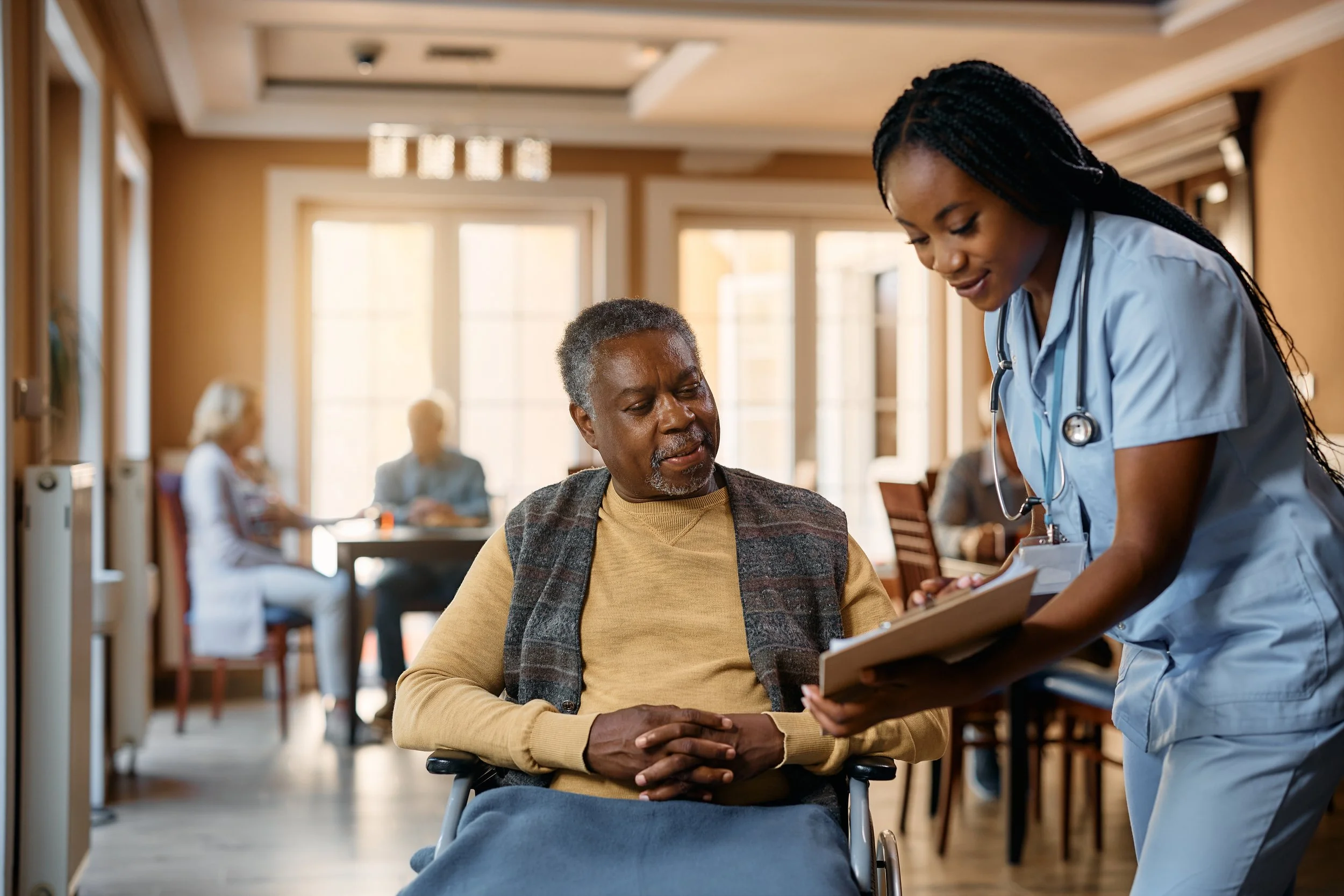 A healthcare professional, a nurse, talking to and taking notes from an elderly patient in a medical facility or pharmacy, with other patients in the background.