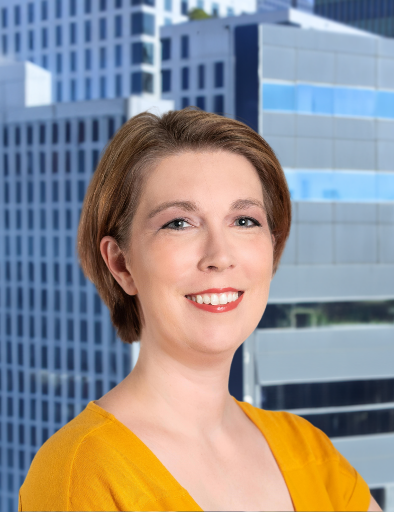 A woman with short brown hair, wearing a yellow top, smiling in front of a high-rise office building in a city.