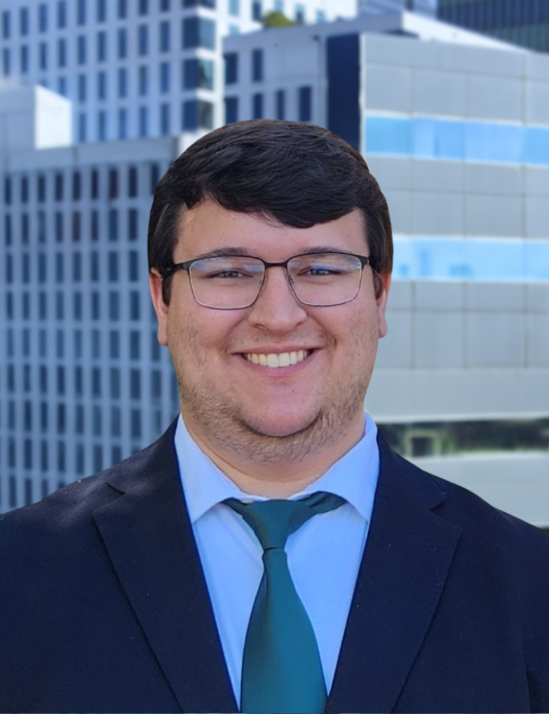 A young man with dark hair and glasses wearing a black suit, light blue dress shirt, and dark green tie. He is smiling and standing outdoors in front of a modern cityscape with tall glass buildings.