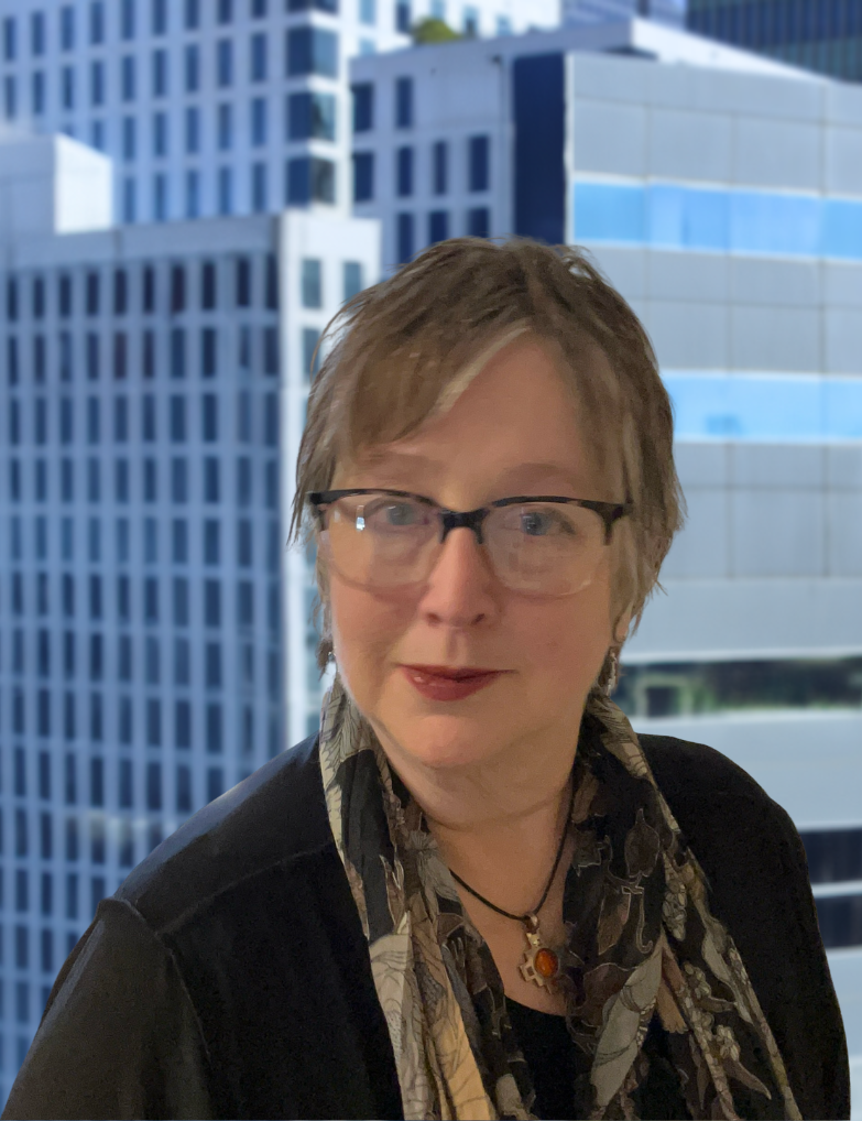 A woman with short, light brown hair and glasses, wearing a black jacket and patterned scarf round her neck, standing in front of a city skyline with high-rise office buildings.