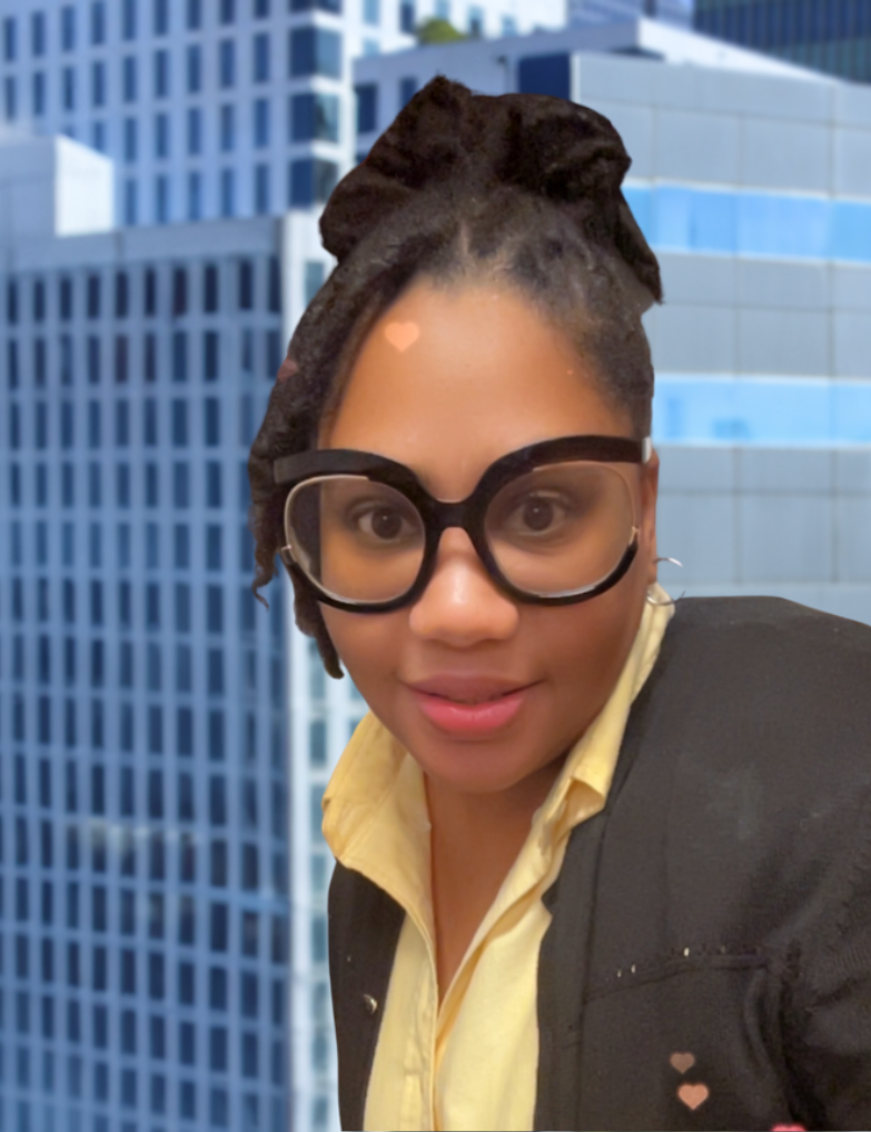 A woman with glasses and styled hair taking a selfie in front of tall office buildings.