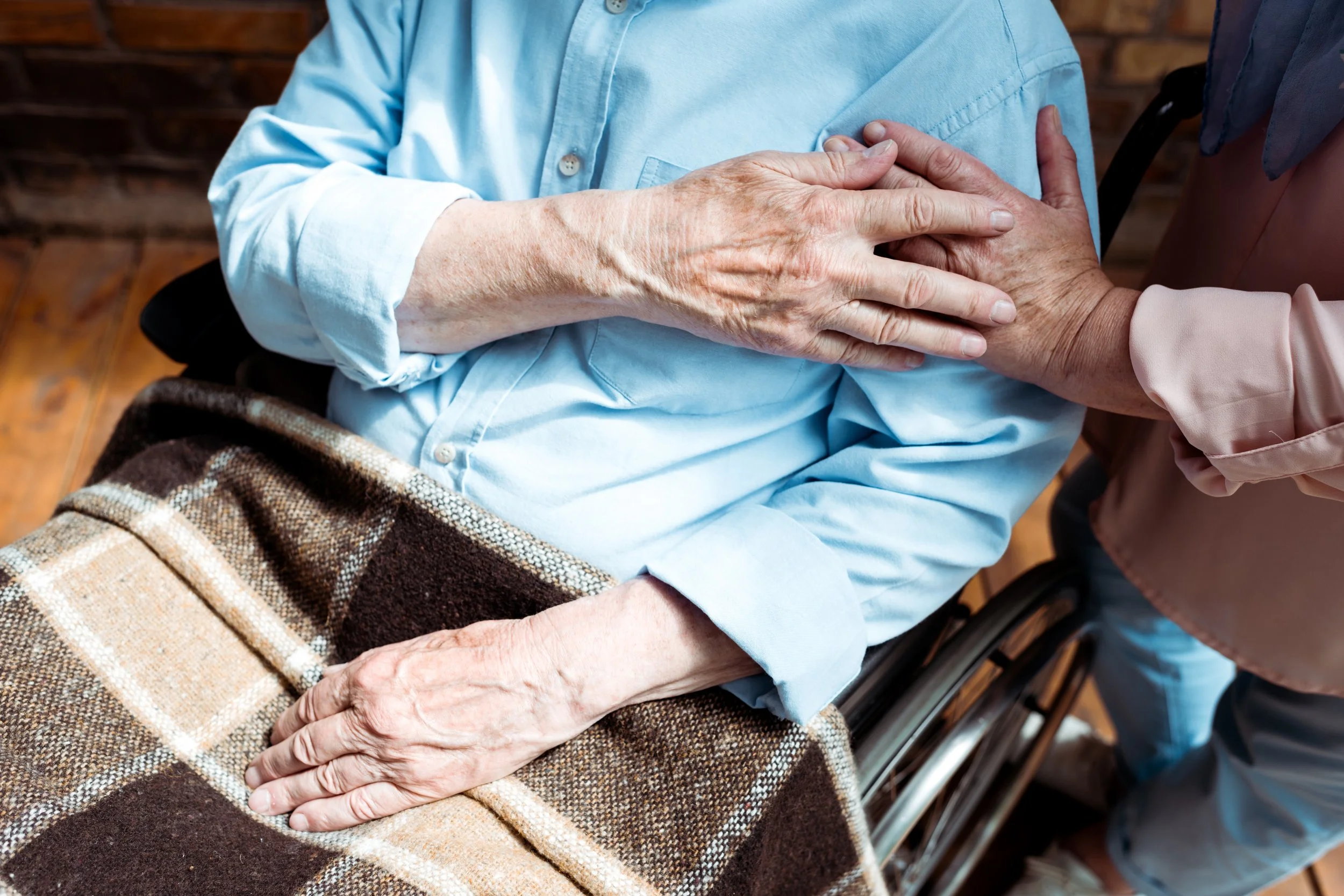 Close-up of an elderly woman in a wheelchair, wearing a light blue shirt, holding hands with another person and showing a gentle touch.