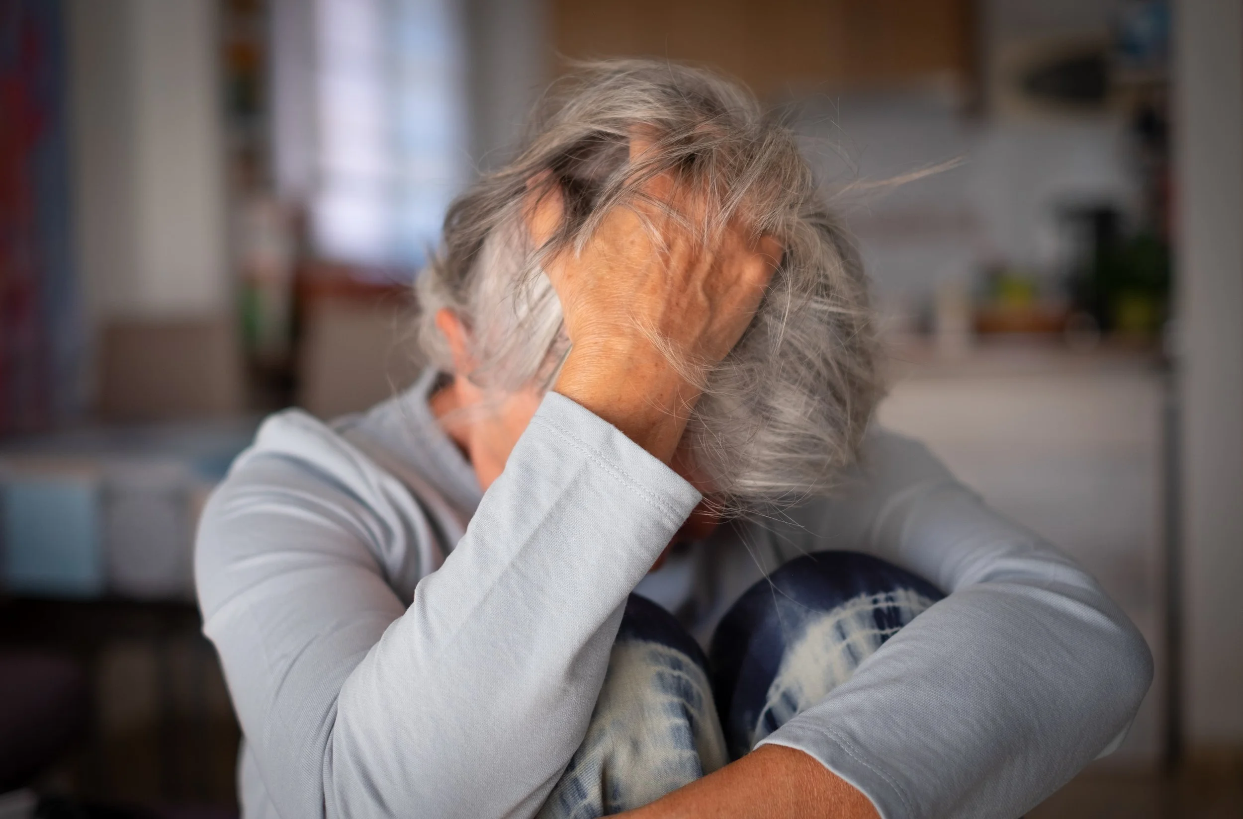 An elderly woman with long gray hair, wearing a light gray long-sleeve shirt, sitting with her elbows on a surface, covering her face with her hands, possibly in distress or sadness, in a home setting.