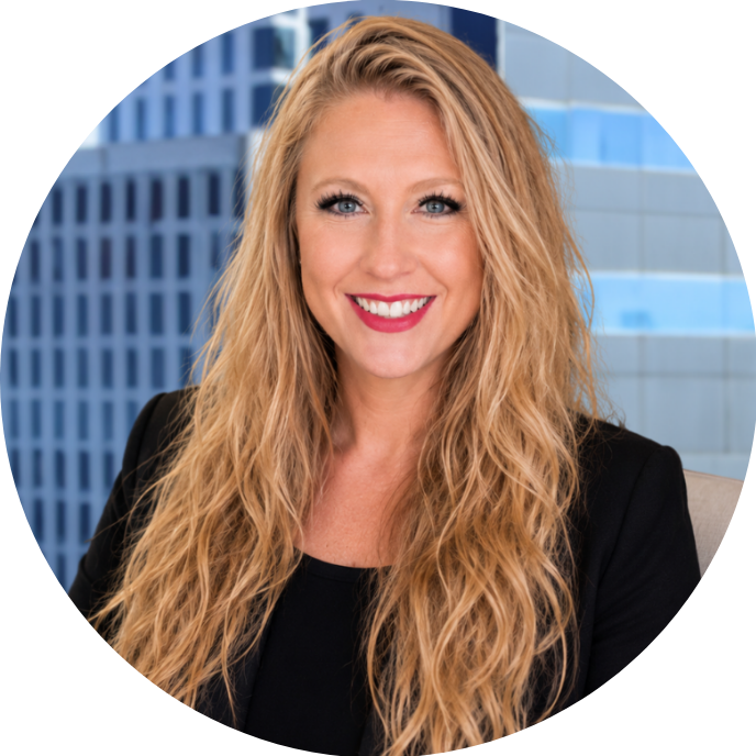 A smiling woman with long, wavy blonde hair and blue eyes, wearing a black blazer, seated in front of a cityscape with tall buildings.