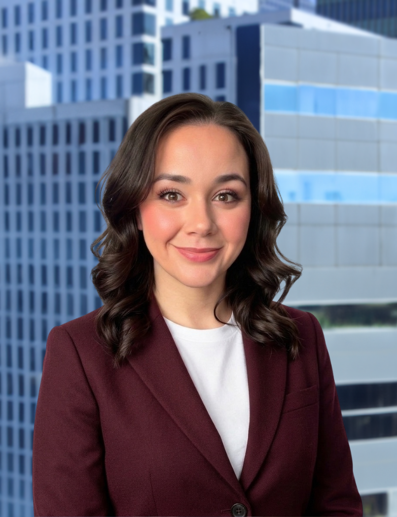 Portrait of a woman with shoulder-length brown hair, wearing a burgundy blazer and white shirt, standing in front of a city office building with glass windows.