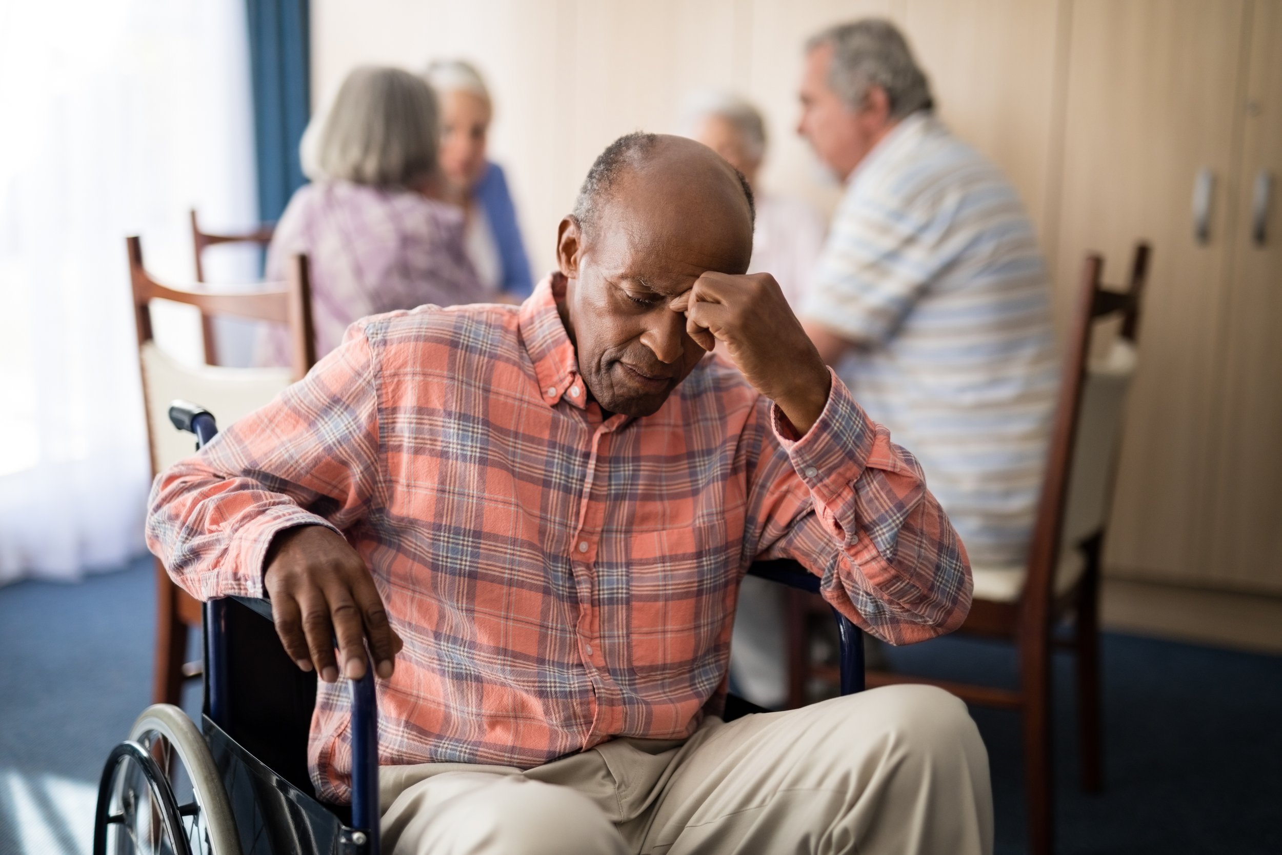 An elderly man in a wheelchair with a distressed expression, holding his forehead, in a room with other elderly people in the background.