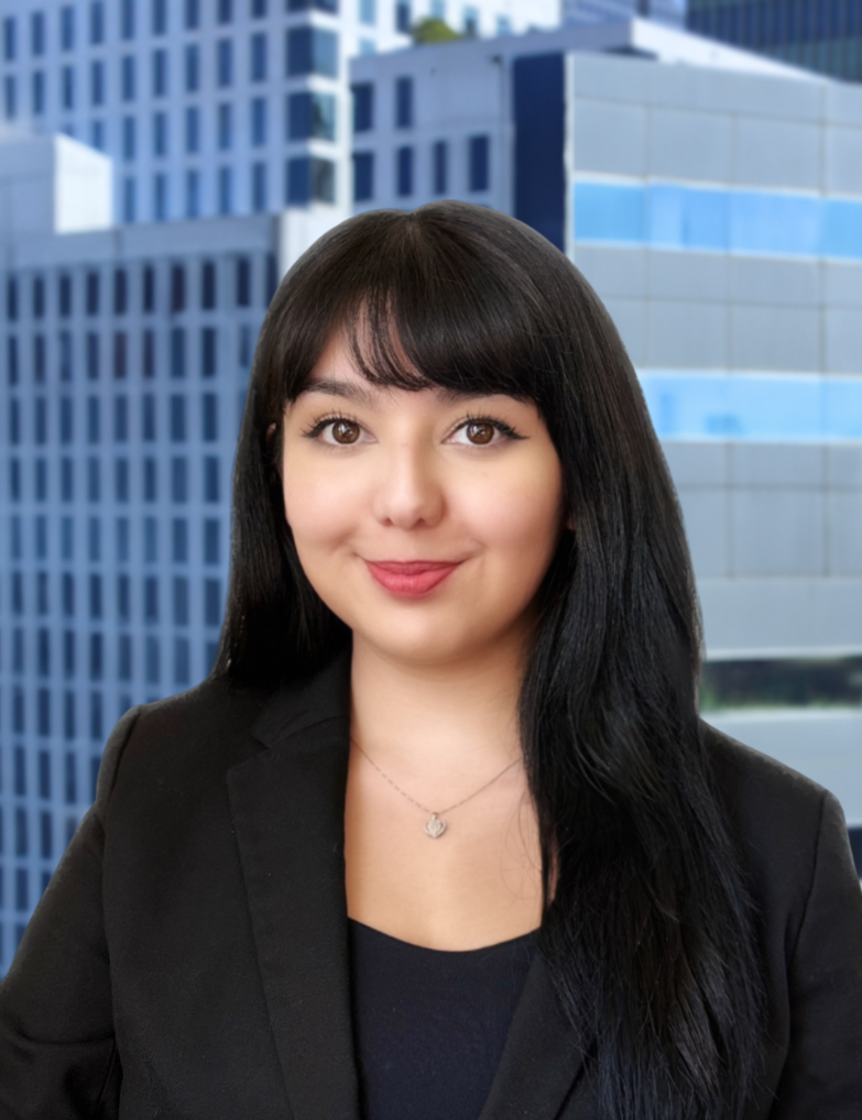 A woman with long black hair and a friendly smile, wearing a black blazer and a necklace, standing in front of a cityscape with tall modern office buildings.