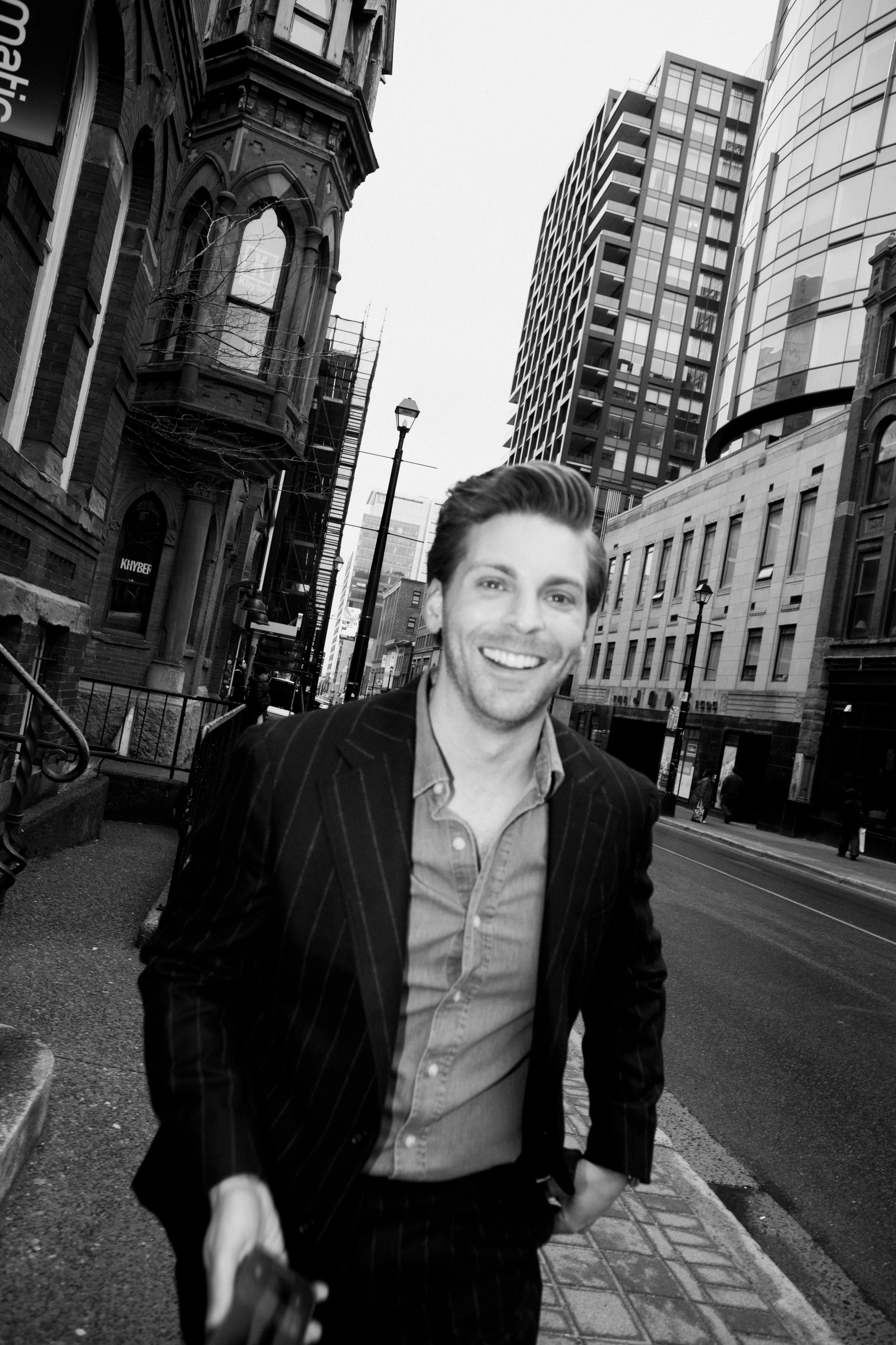 A smiling man in a suit and open-collared shirt walking on a city sidewalk with tall modern and historic buildings in the background, in black and white.