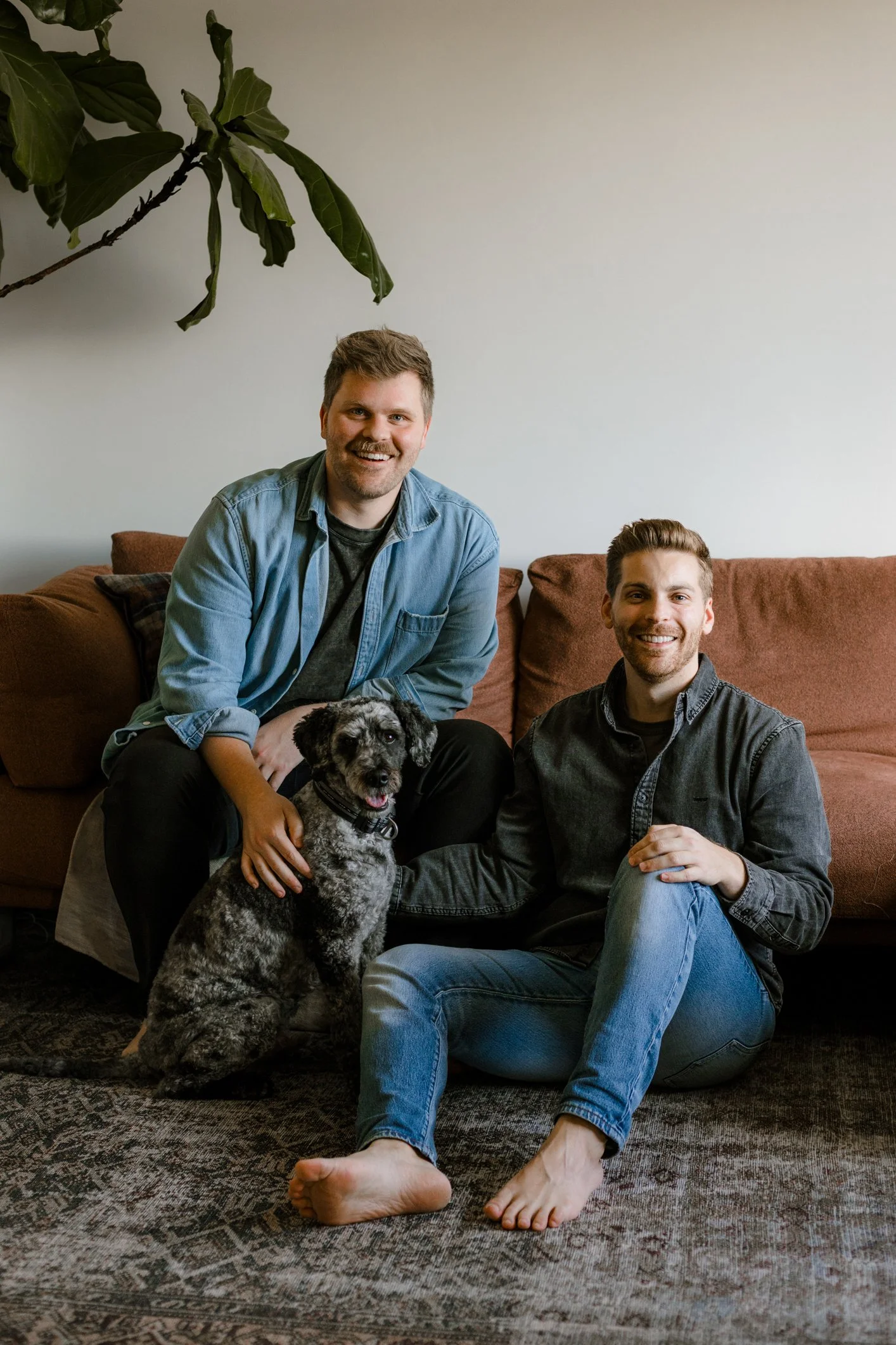 Two smiling men sitting on the floor in front of a sofa, with a large dog between them. One man is sitting on the floor with his legs crossed, and the other is sitting on the floor with his knees up and feet touching the ground. They are inside a living room with a white wall and a large plant hanging from the ceiling.
