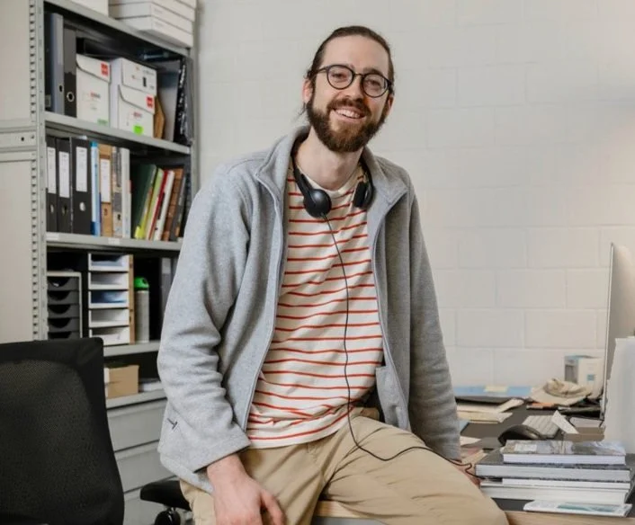 Junger Mann mit Brille, Bart, grauer Jacke und Kopfhörern sitzt an einem Schreibtisch in einem Büro mit Büchern und Aktenregal im Hintergrund.