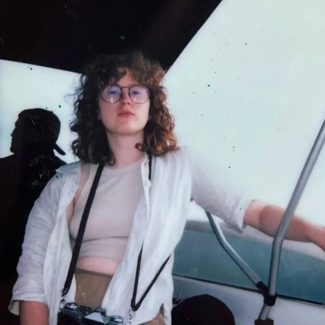 Junge Frau mit Brille, lockigen Haaren, weiße Jacke und Kamera um den Hals, sitzt in einem öffentlichen Verkehrsmittel.
