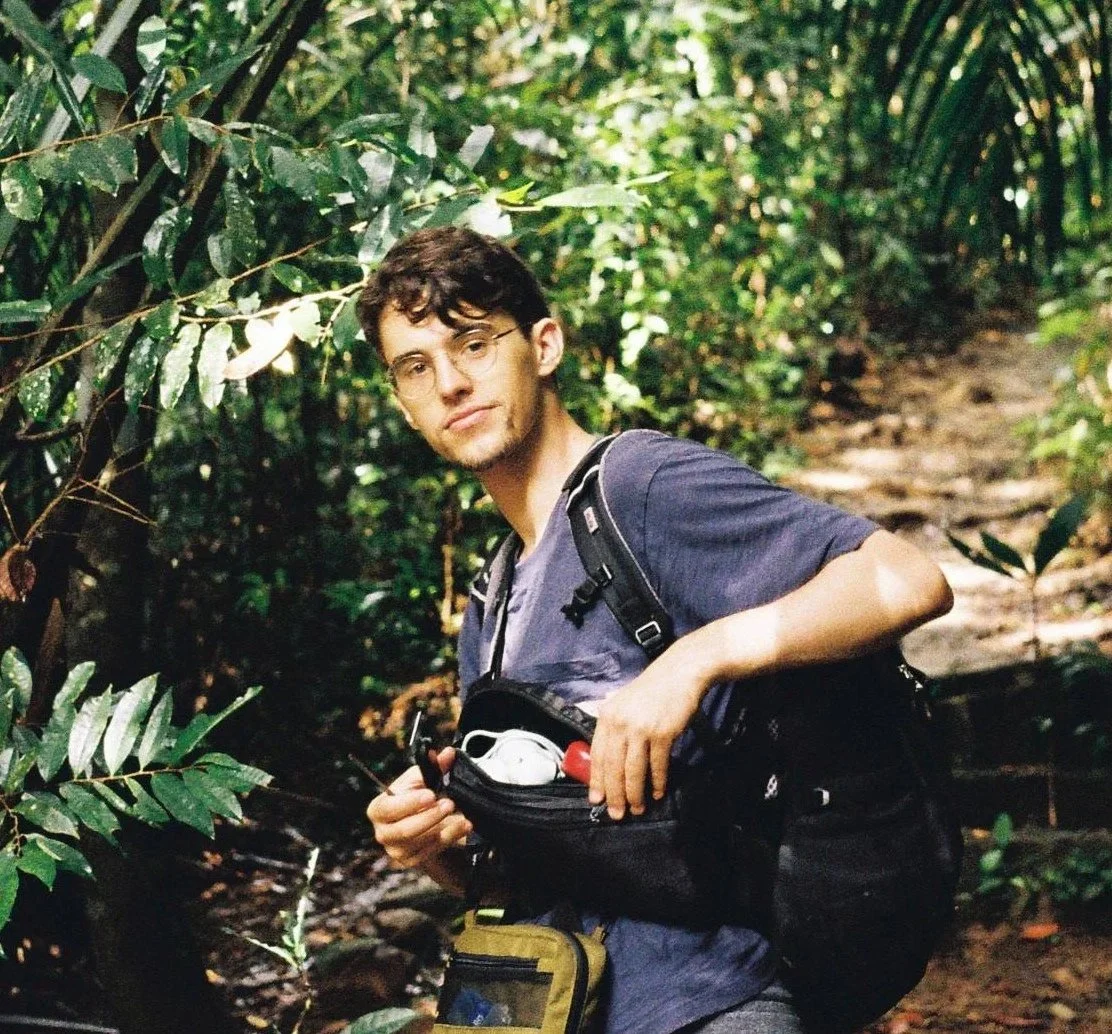 Ein junger Mann mit Brille, dunkelwogigem Haar und einem Rucksack, der in einem Wald steht, mit einer kleinen Gürteltasche um die Taille.