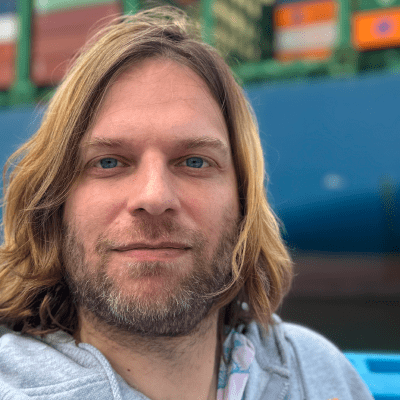 Nahaufnahme eines Mannes mit langen braunen Haaren und Bart, trägt graues Hoodie, im Hintergrund eine blaue Fähre oder ein Boot