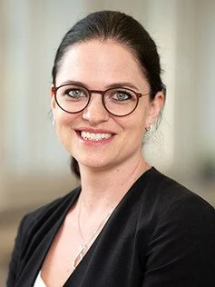 Porträt einer lachenden Frau mit dunklen Haaren, Brille und dunklem Business-Kleidung, vor einem hellen Hintergrund.