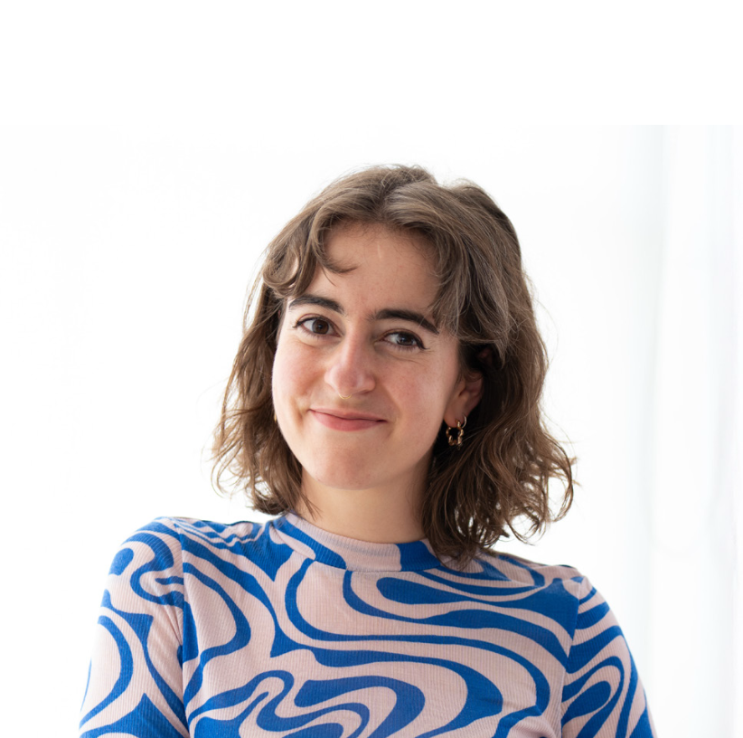 Junge Frau mit braunen, lockigen Haaren und einem blauen und beige Muster-Shirt, die lächelnd in die Kamera schaut in einem hellen Raum.