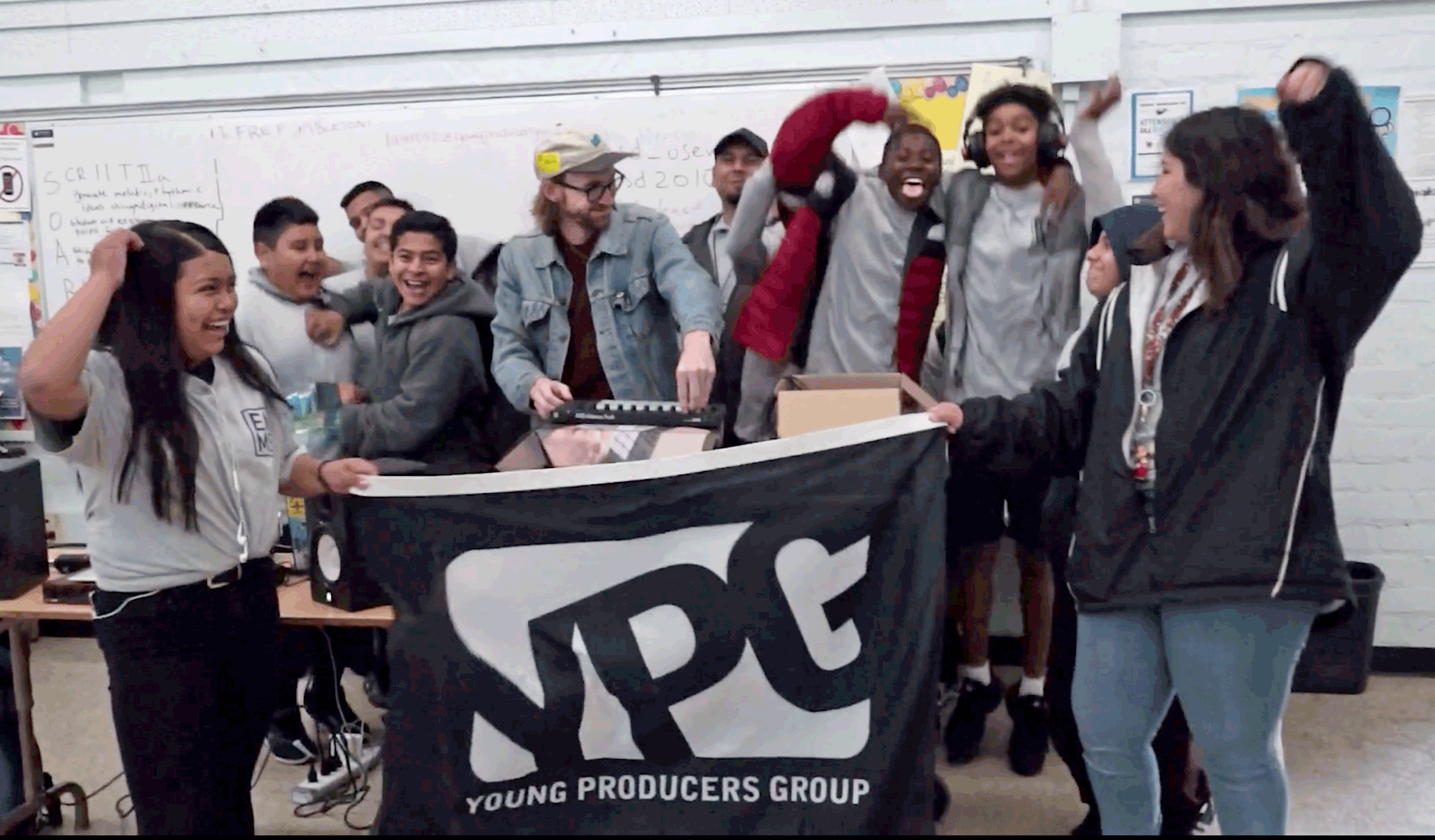 Students and a teacher celebrate during a Young Producers Group Listening Party, holding a YPG flag.