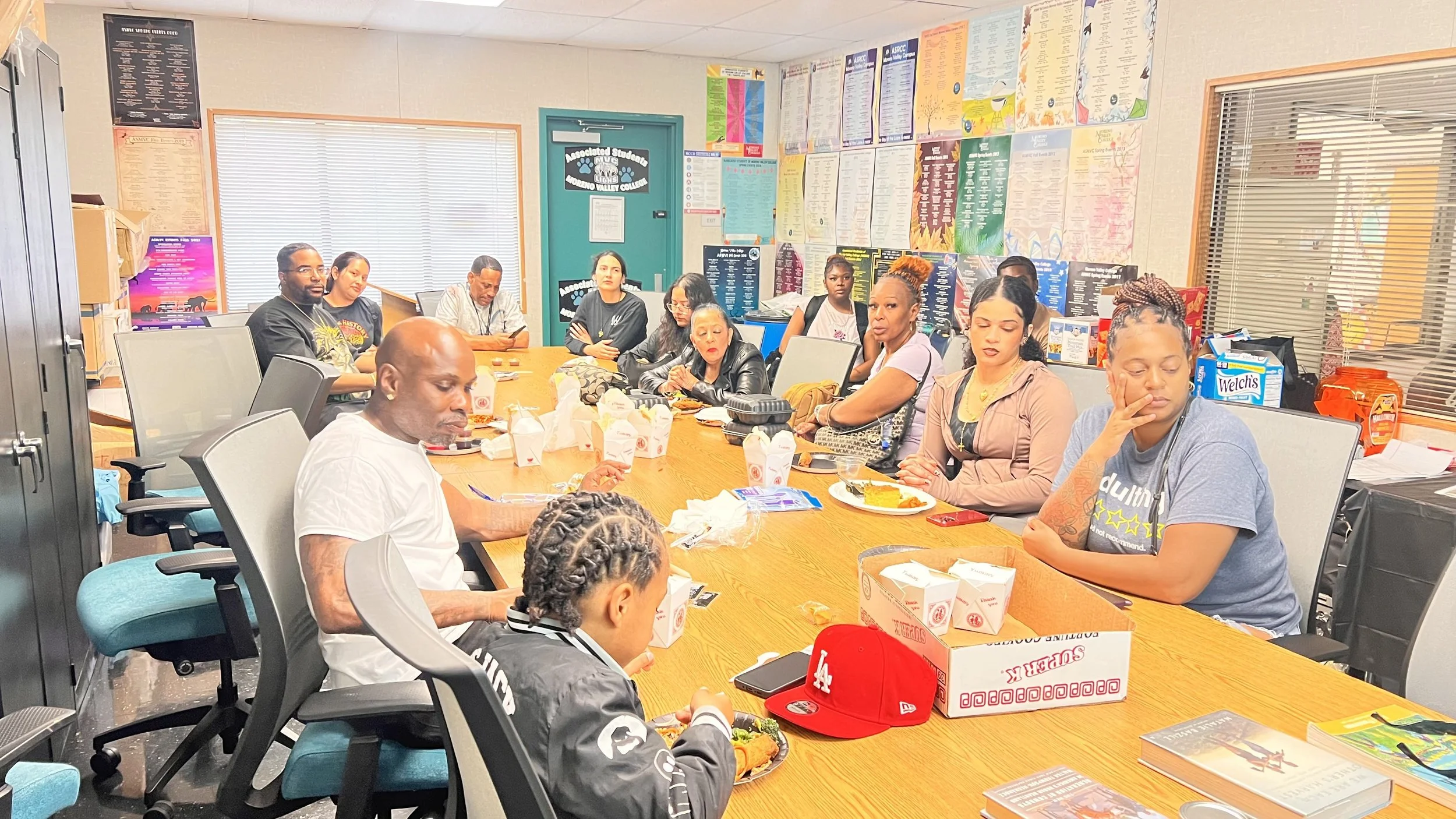 Group of people sitting around a large conference table in a classroom or meeting room, with snacks, drinks, and food containers on the table, and colorful posters on the walls.