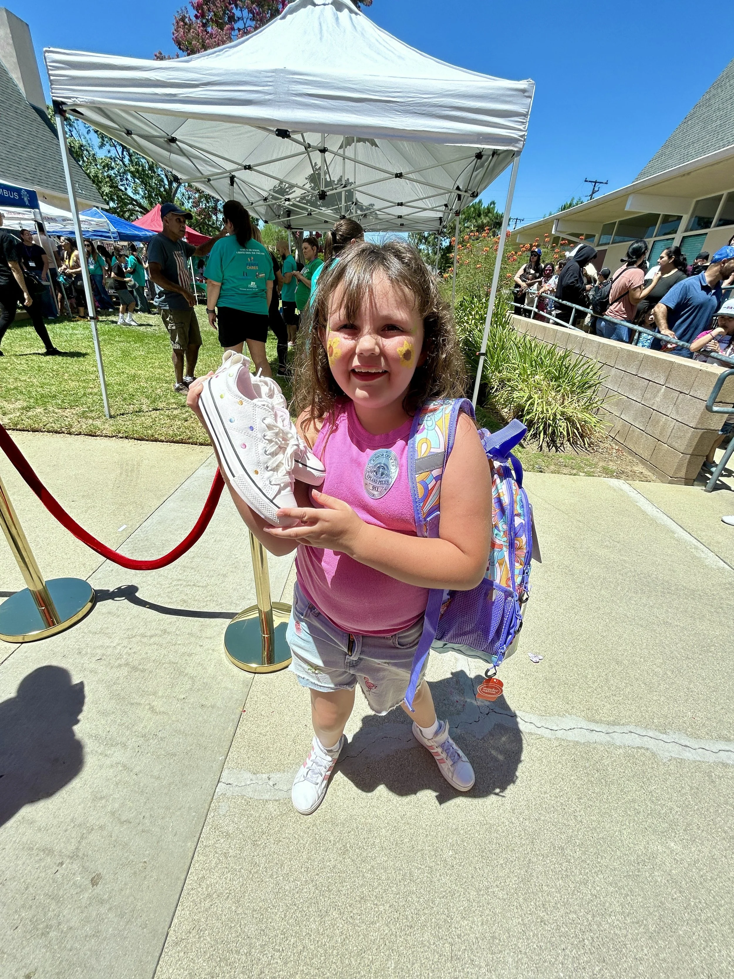 Children and adults standing outdoors in a park or playground area, with a person dressed in a brown bear costume wearing a white cape that reads 'Good Neil Bear.' The children are wearing backpacks and are gathered around listening to a speaker. There are trees, houses, and a playground structure in the background.