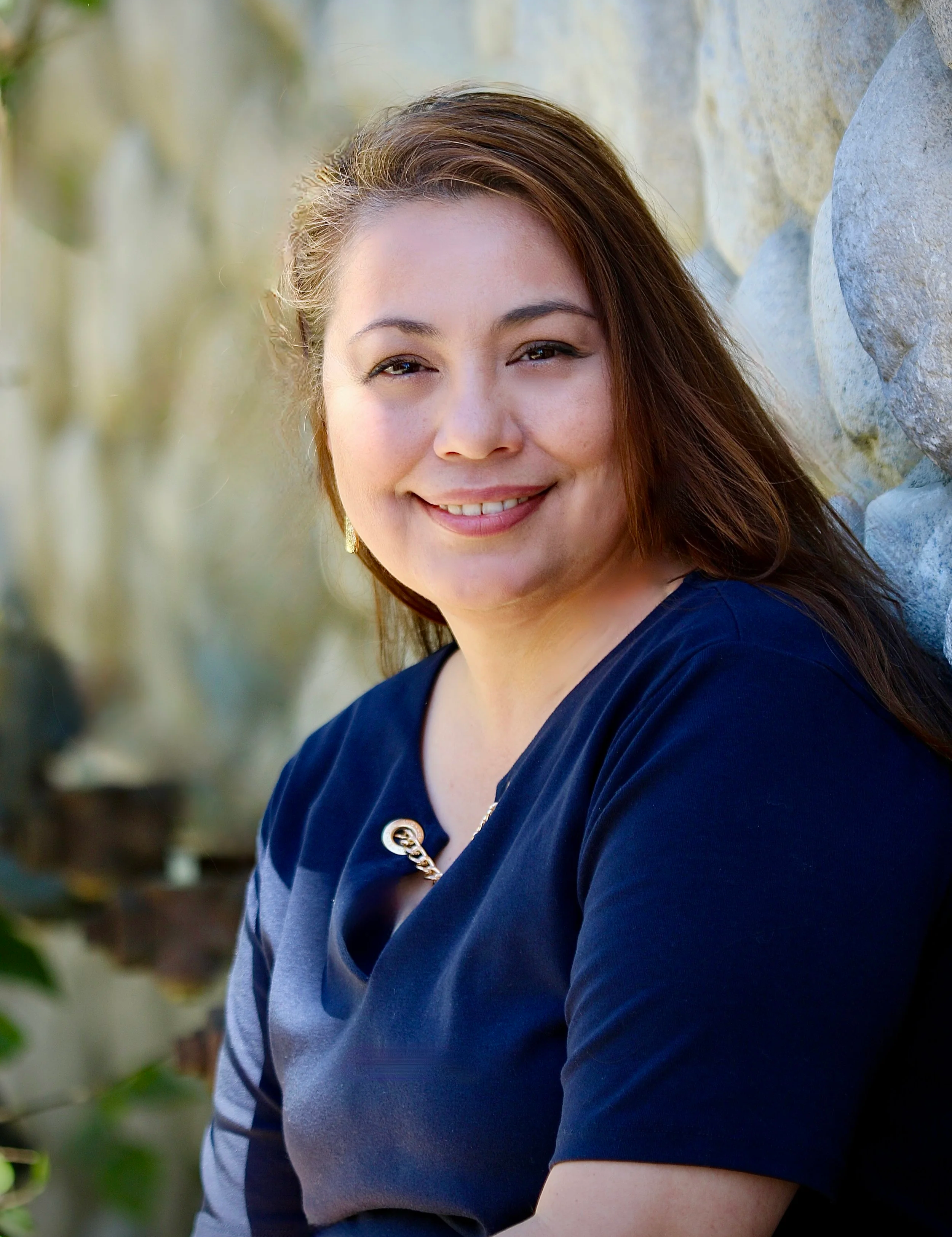 A woman with long, wavy brown hair smiling outdoors, wearing a black top with pleats and a name tag that says Linda Contreras, standing in front of a tree with green leaves and pink flowers.