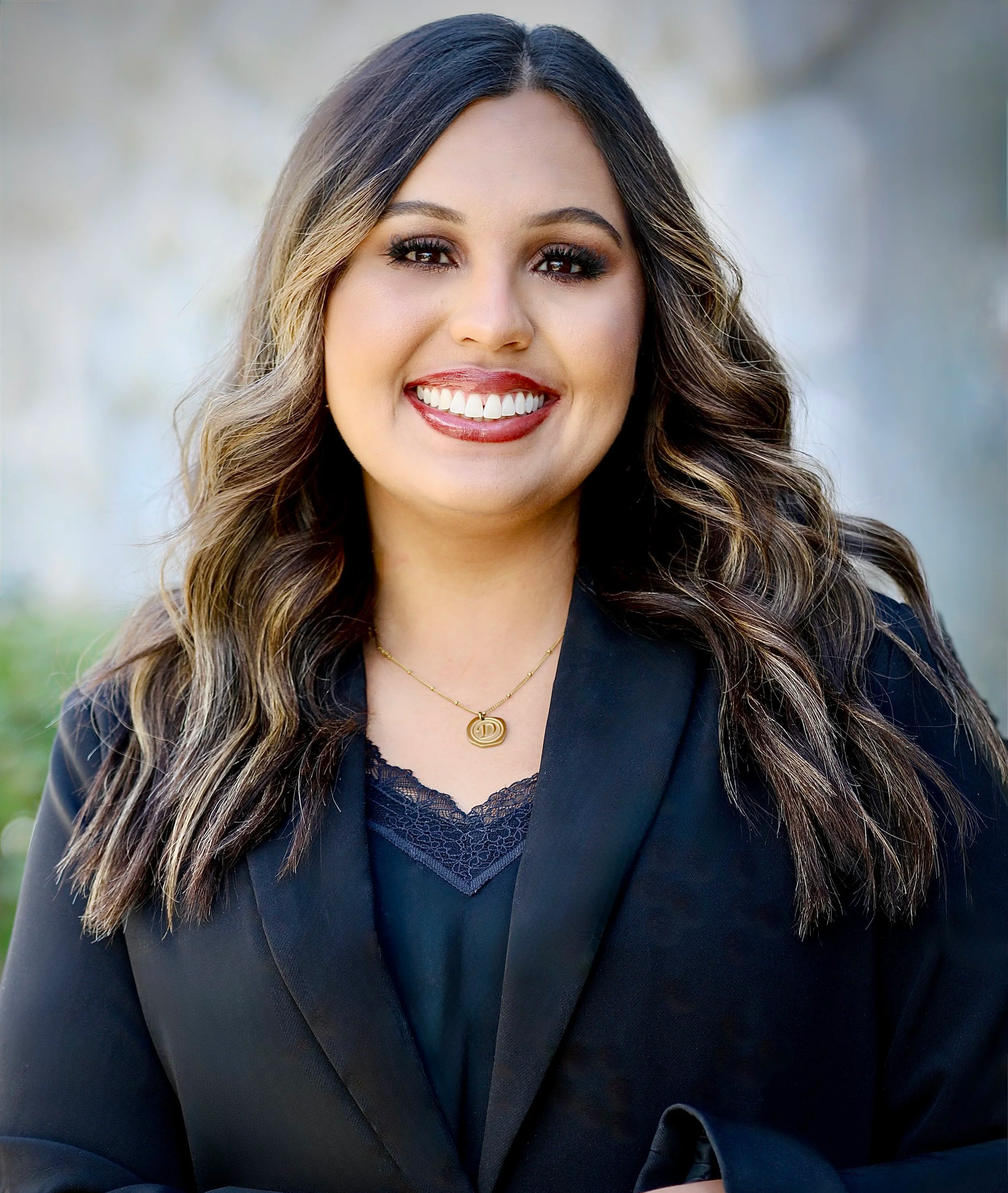 A woman with long dark hair smiling outdoors, wearing a black top and a gold necklace with a circular pendant, standing in front of a tree with green leaves.