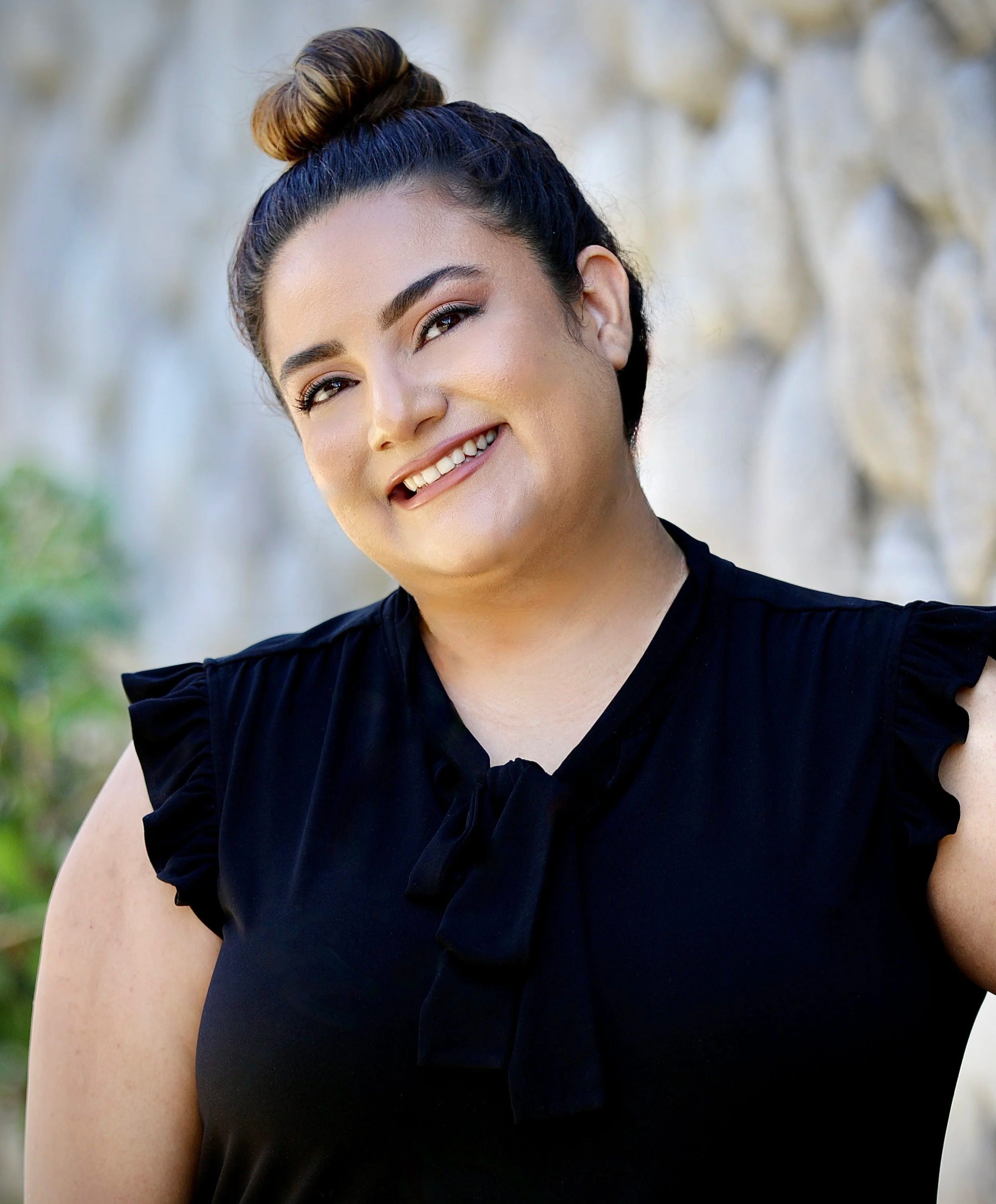 A woman with long, wavy brown hair wearing a black shirt and a name badge outdoors with trees and a building in the background.