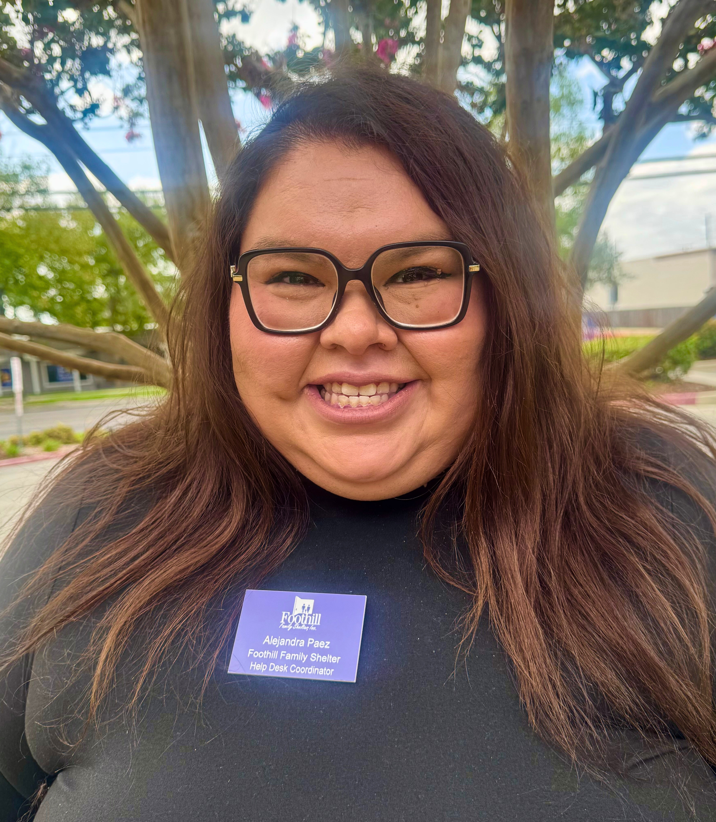 A woman with long brown hair, glasses, and a black shirt standing outdoors in front of a tree with pink flowers. She is smiling and wearing a blue name tag that reads 'Alejandra Paez, Foothill Family Shelter, Help Desk Coordinator.'