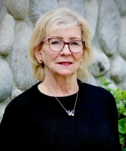 A woman wearing glasses, a black top, butterfly necklace, and earrings, smiling outdoors, with trees and a parking lot in the background, wearing a name tag that reads 'Bonnie Cole, Foothill Family Shelter, Interim Executive Director'.