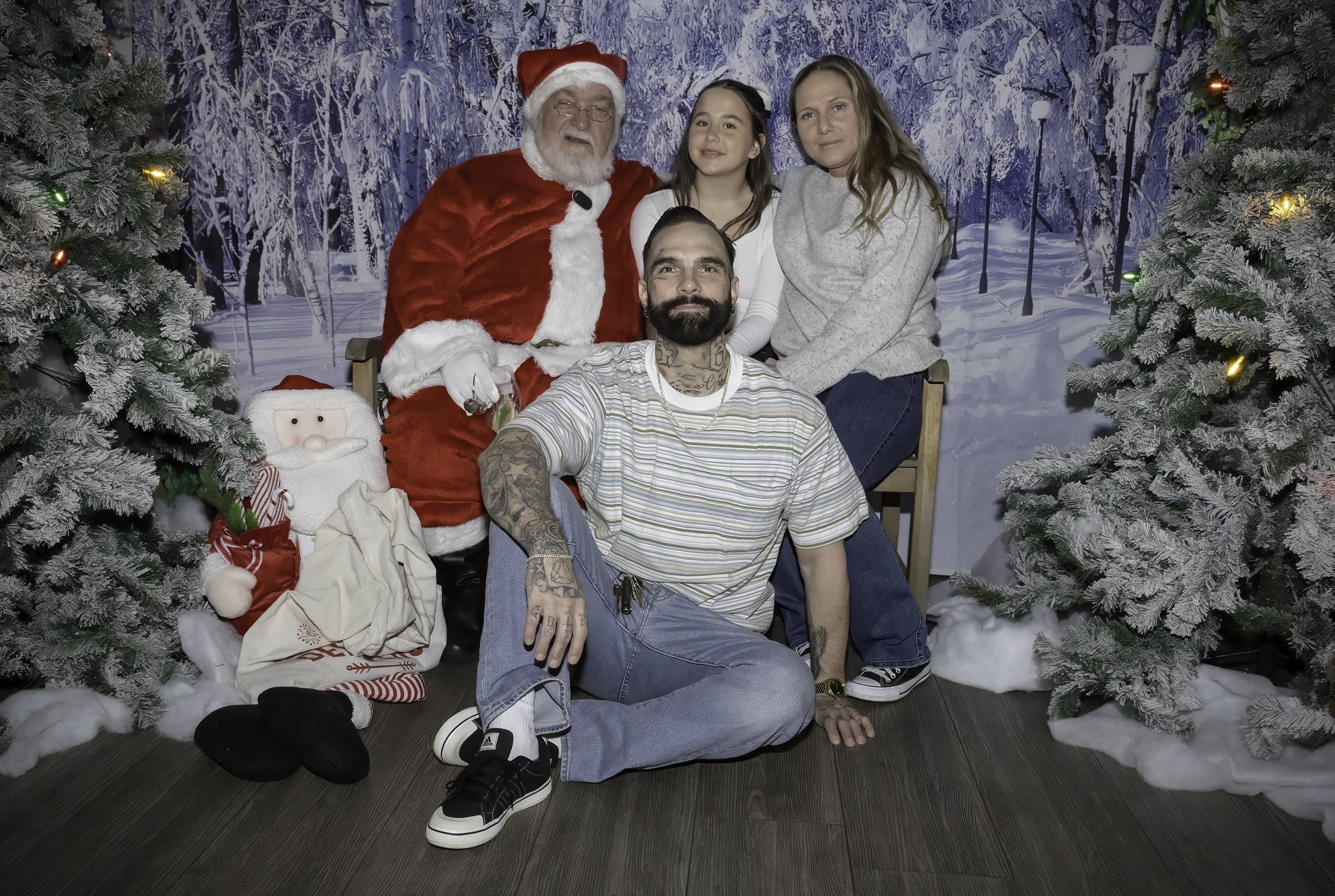 Group of children and adults gathered around Santa Claus in front of a winter-themed backdrop with snow-covered trees and sky, decorated for Christmas celebration.
