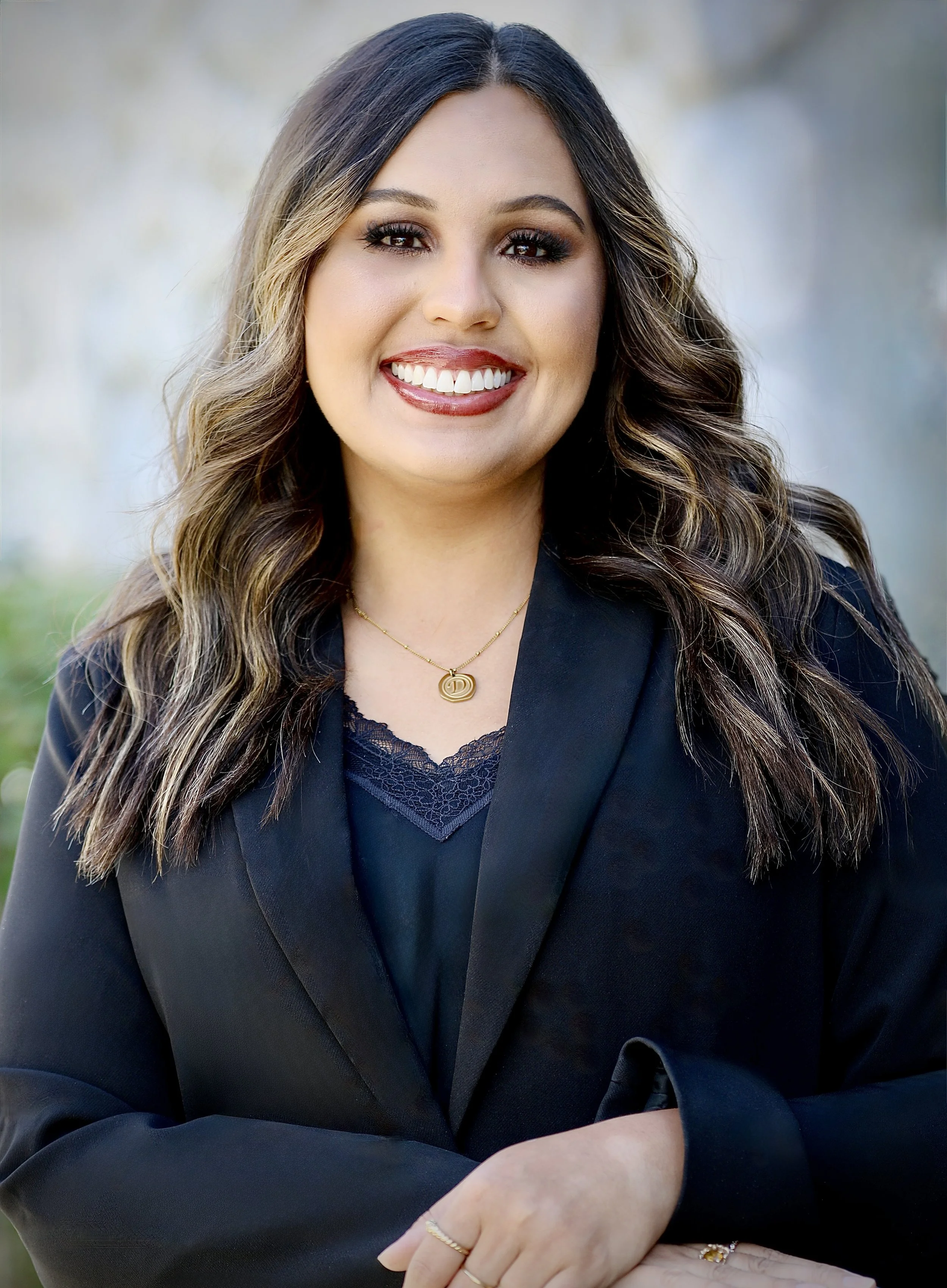 A woman with long dark hair smiling outdoors, wearing a black top and a gold necklace with a circular pendant, standing in front of a tree with green leaves.