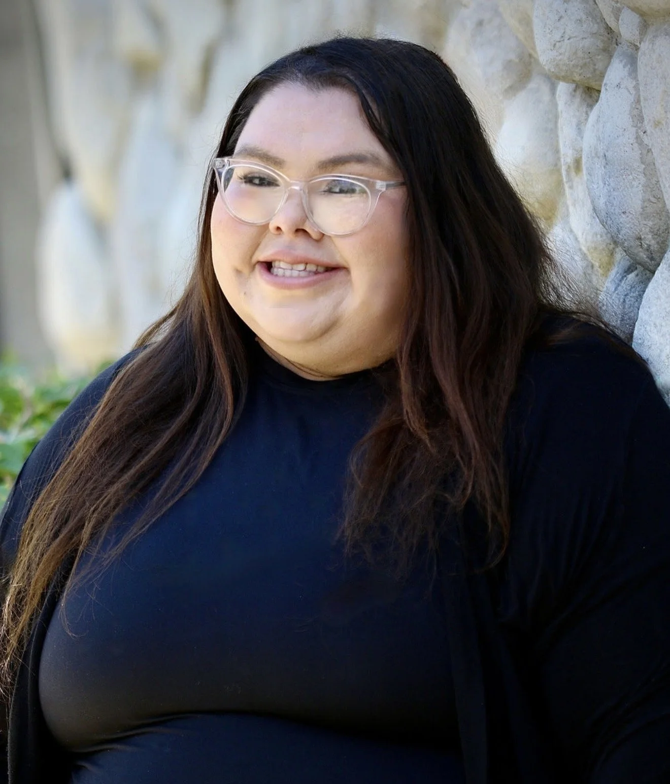 A woman with long brown hair, glasses, and a black shirt standing outdoors in front of a tree with pink flowers. She is smiling and wearing a blue name tag that reads 'Alejandra Paez, Foothill Family Shelter, Help Desk Coordinator.'