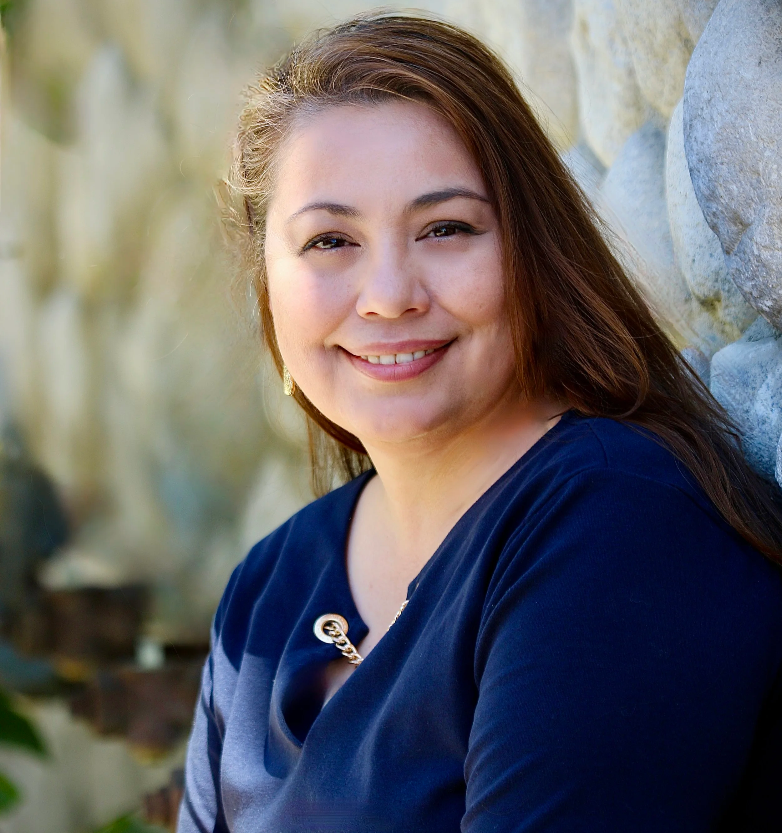 A woman with long, wavy brown hair smiling outdoors, wearing a black top with pleats and a name tag that says Linda Contreras, standing in front of a tree with green leaves and pink flowers.