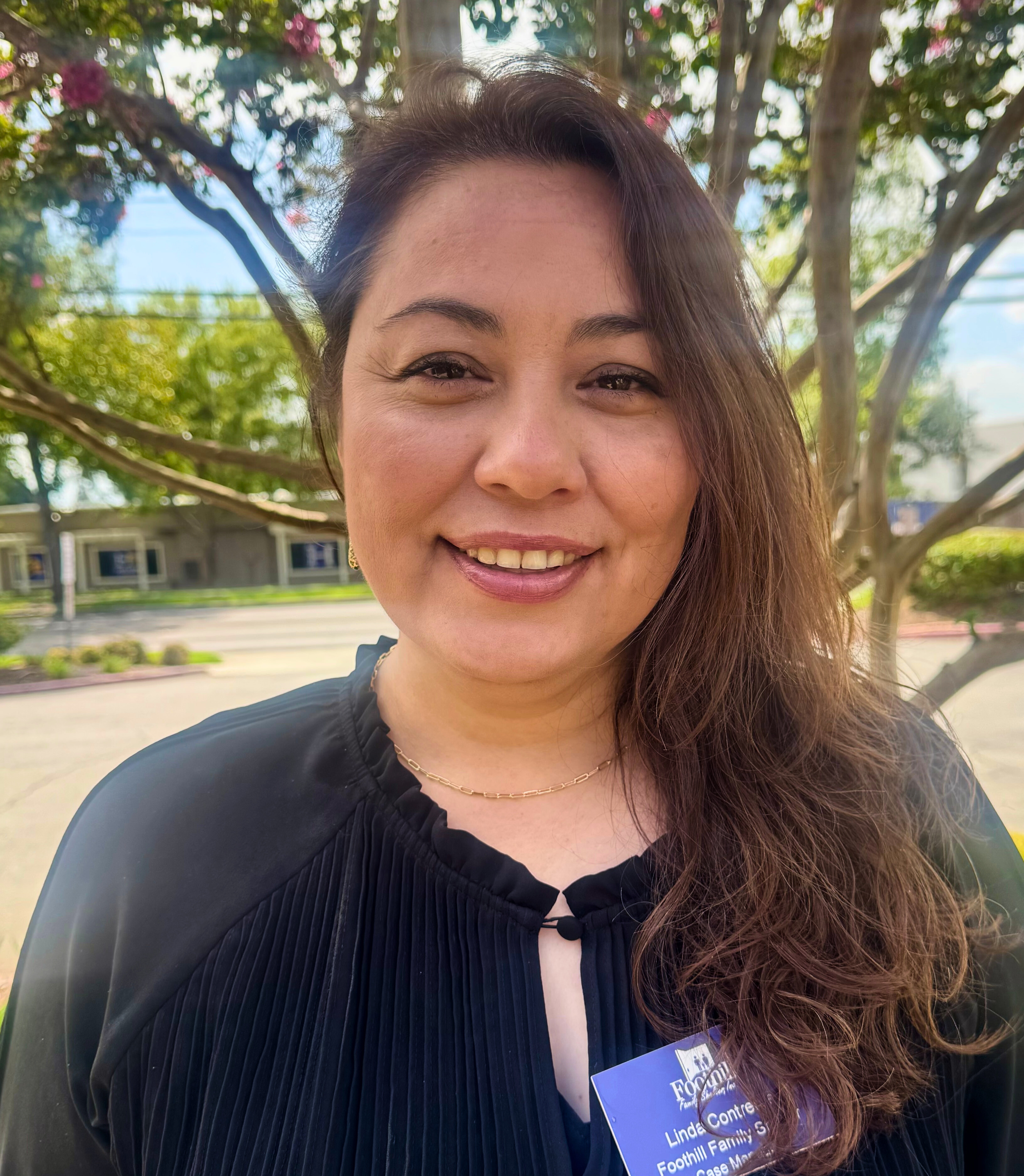 A woman with long, wavy brown hair smiling outdoors, wearing a black top with pleats and a name tag that says Linda Contreras, standing in front of a tree with green leaves and pink flowers.