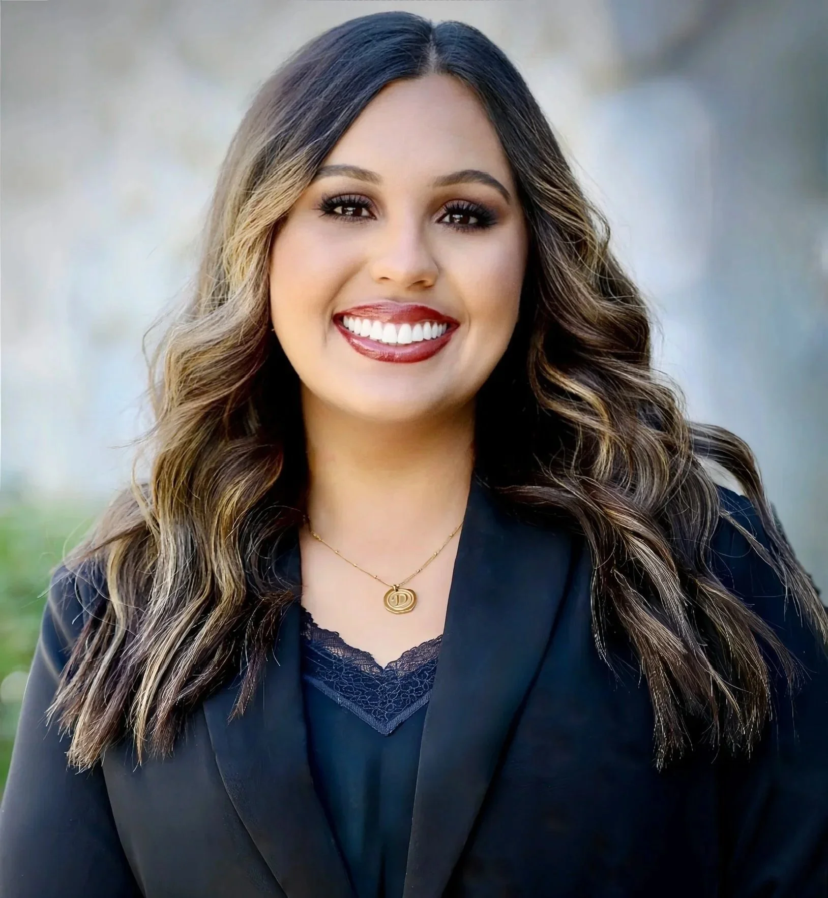 A woman with long dark hair smiling outdoors, wearing a black top and a gold necklace with a circular pendant, standing in front of a tree with green leaves.