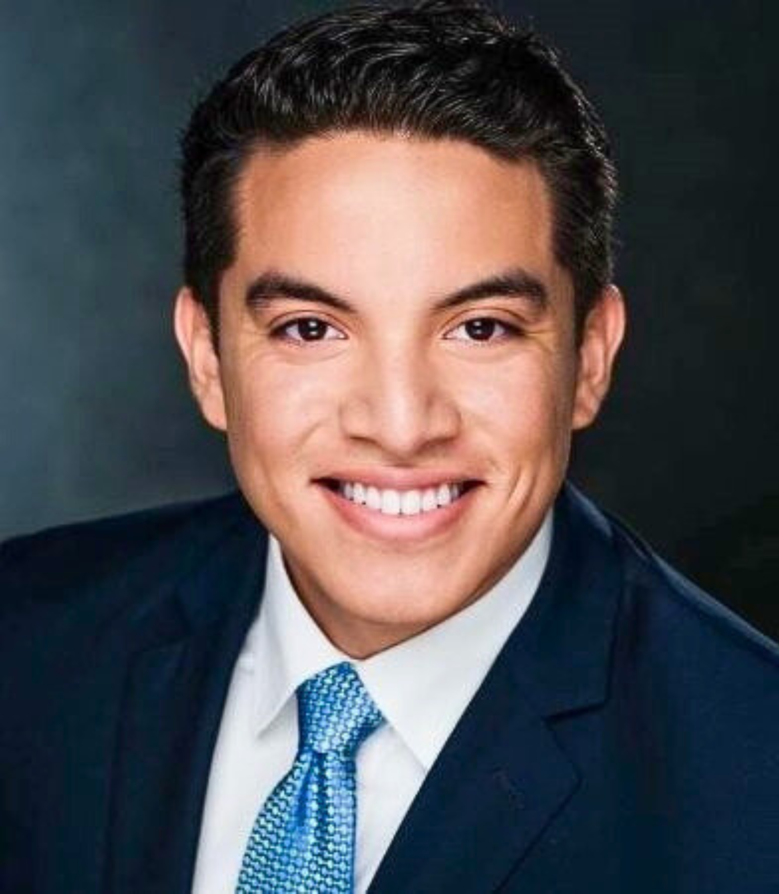 A young man wearing a dark suit, white shirt, and blue tie, smiling at the camera against a dark background.