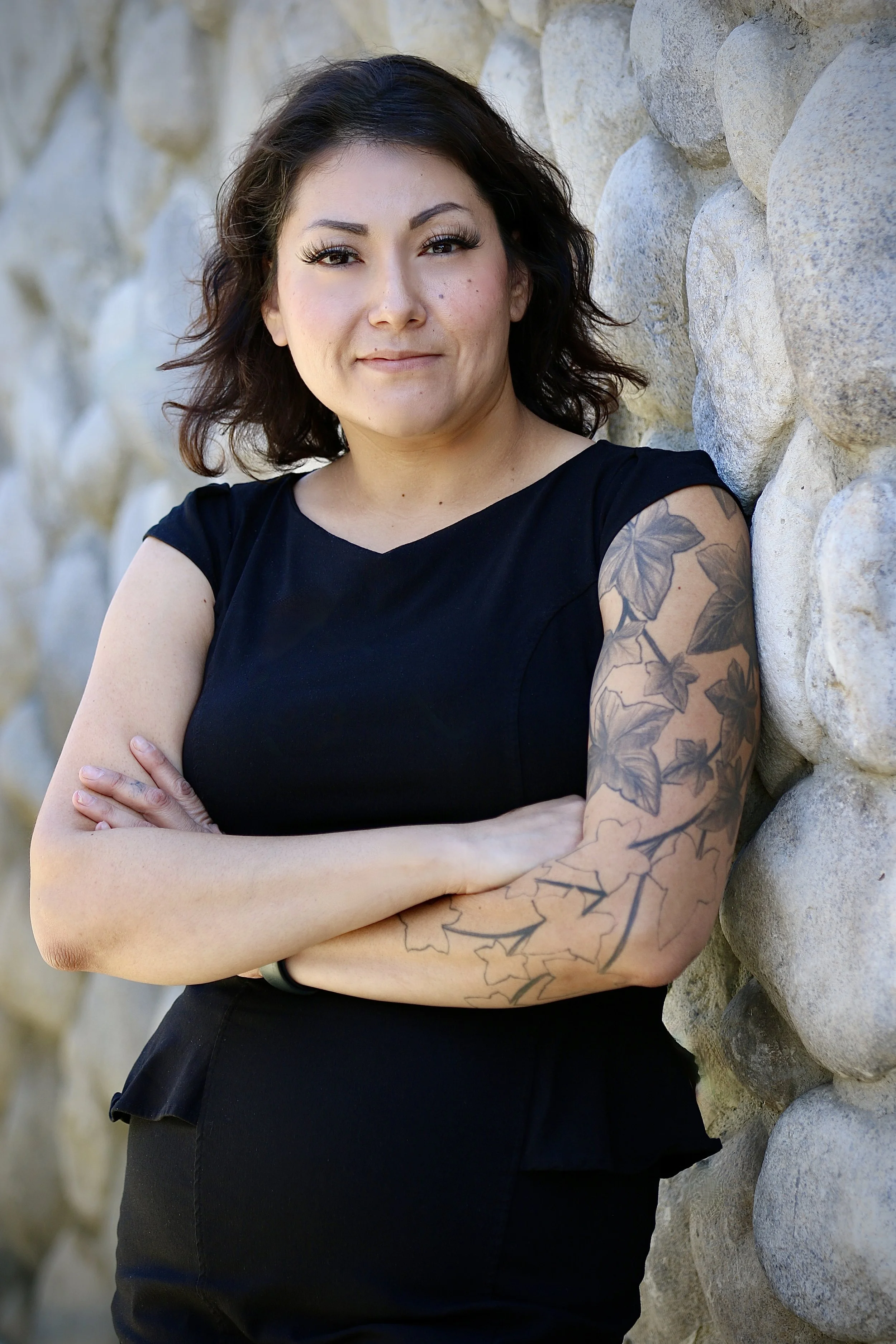 A woman with short dark hair wearing a black turtleneck and a name tag, standing outdoors near a tree, with a street and some buildings in the background.