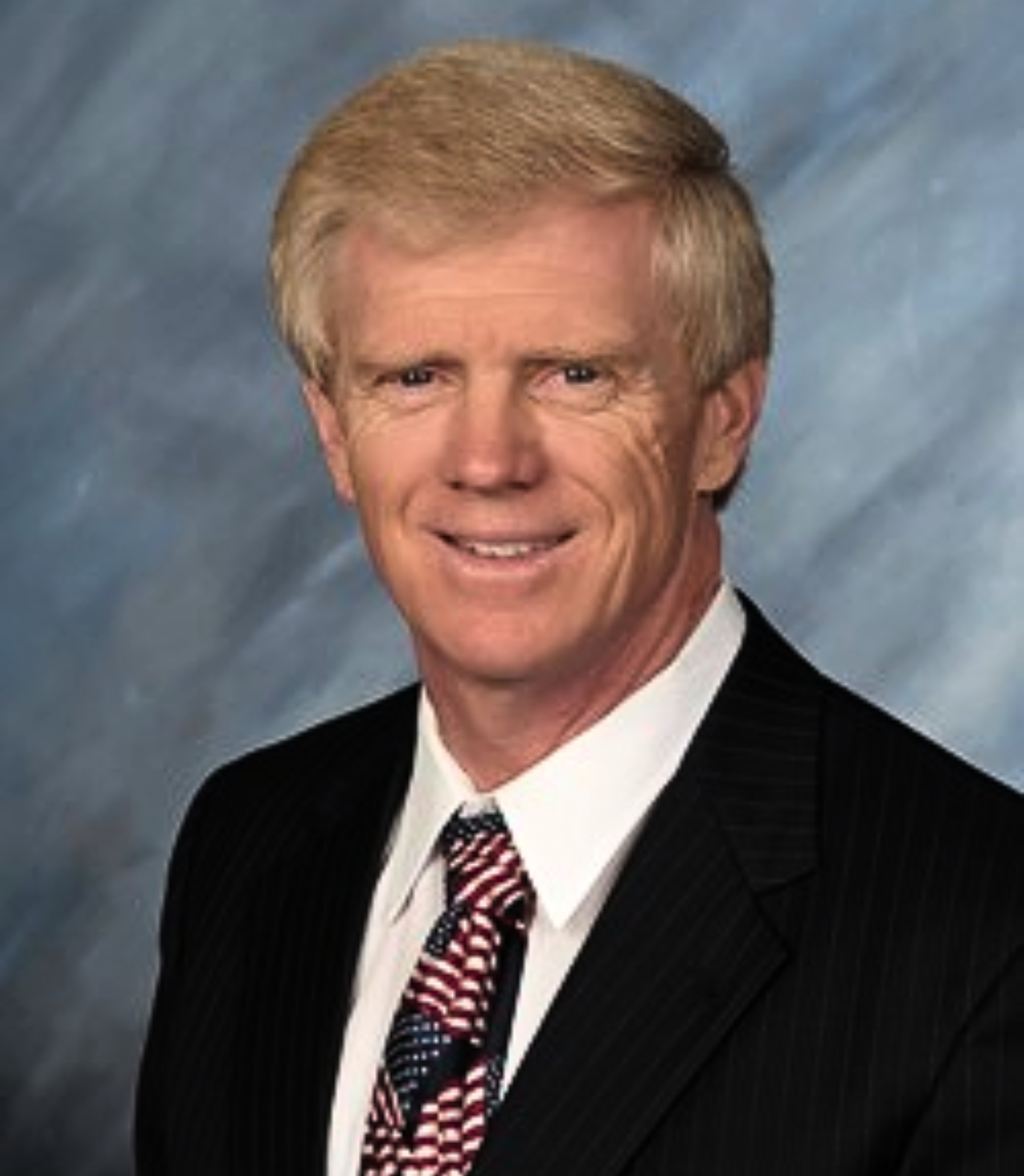 A smiling man in a blue suit, white shirt, and a plaid red and blue tie, posing for a formal portrait with a cloudy background.