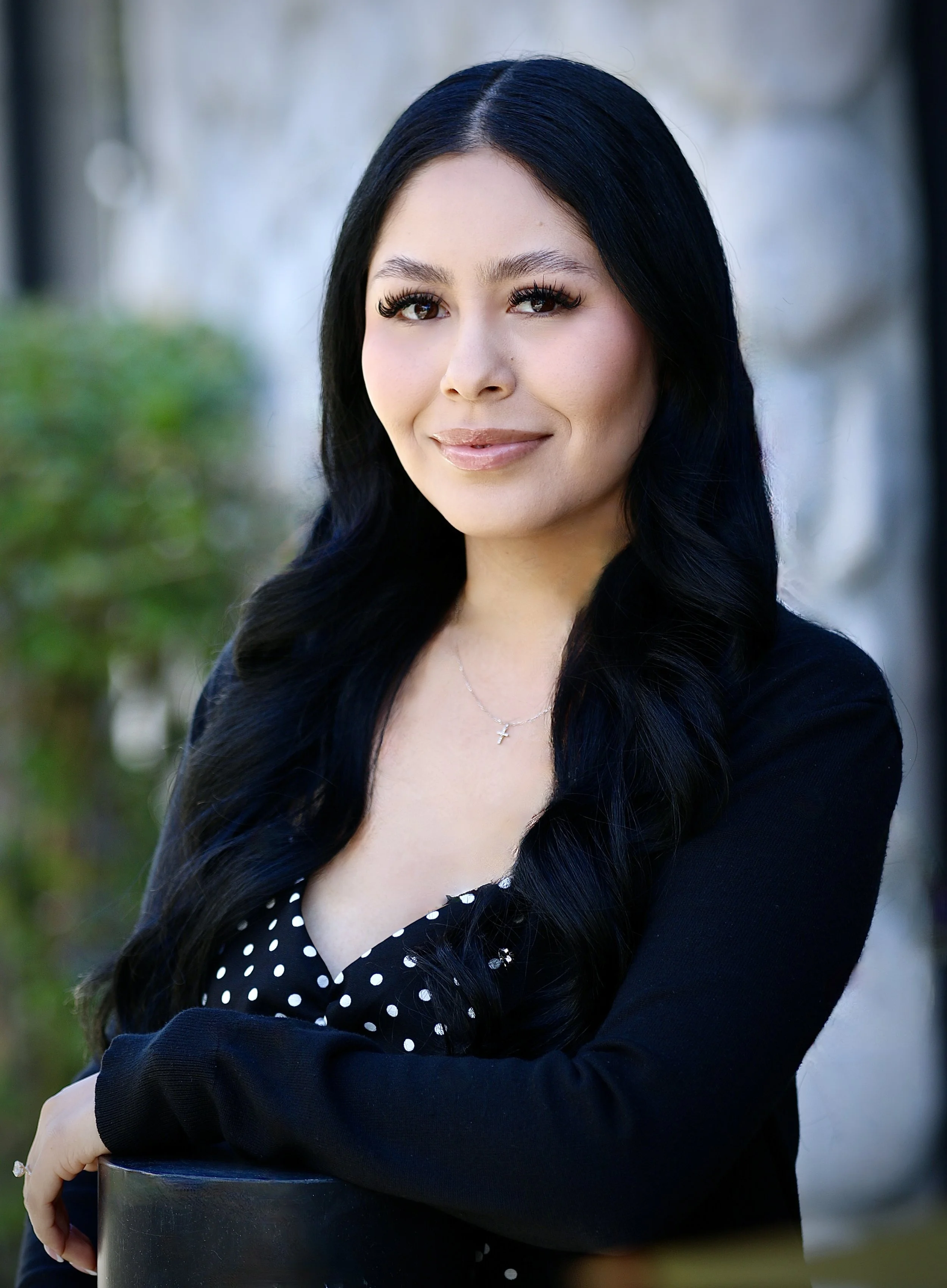 A woman with long black hair wearing a black sleeveless top and a name badge standing outdoors in front of a tree with green foliage and buildings in the background.
