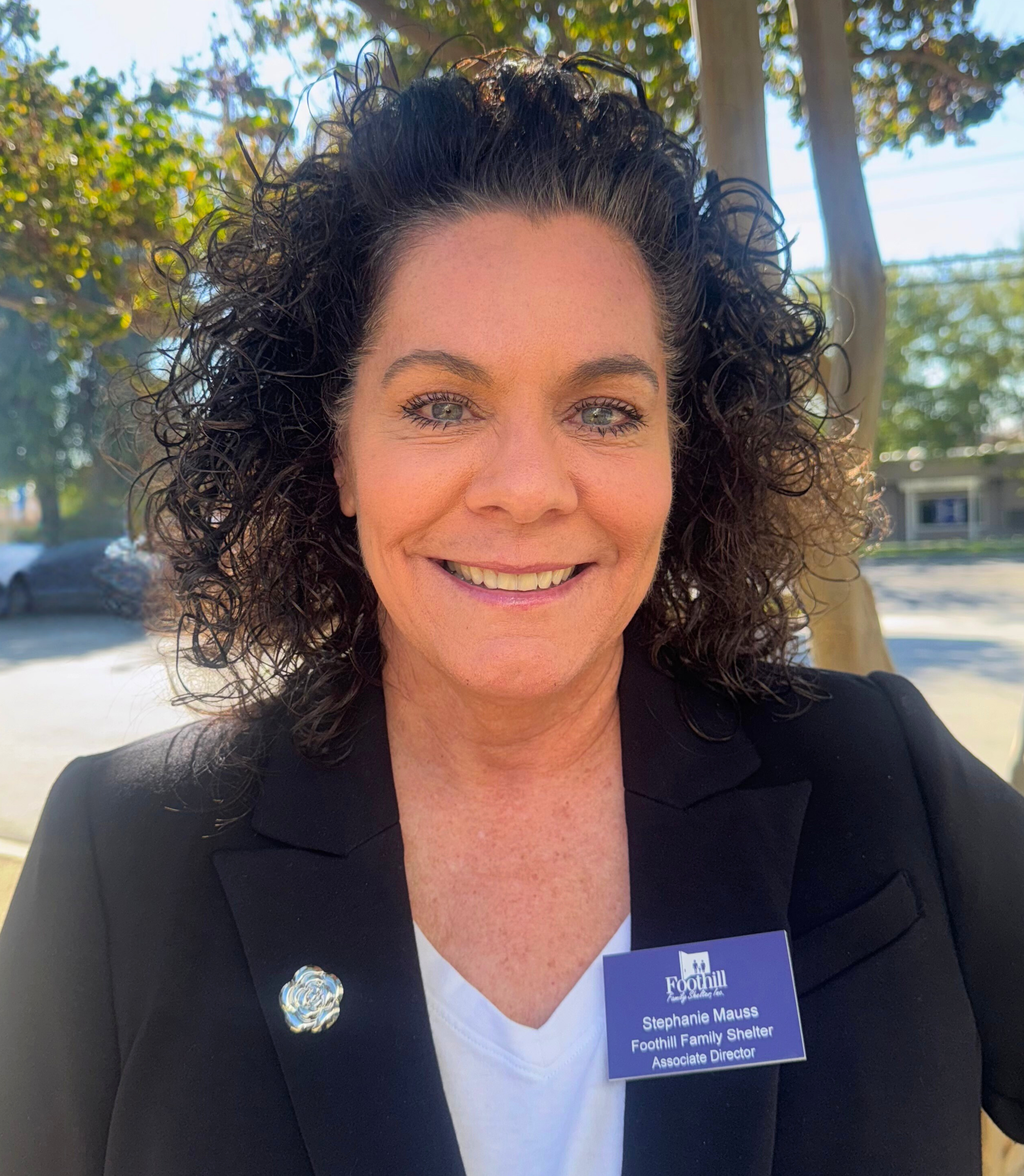 A woman with curly dark hair smiling outdoors, wearing a black blazer with a name tag that reads 'Stephanie Mauss, Foothill Family Shelter, Associate Director', and a decorative pin on her blazer.