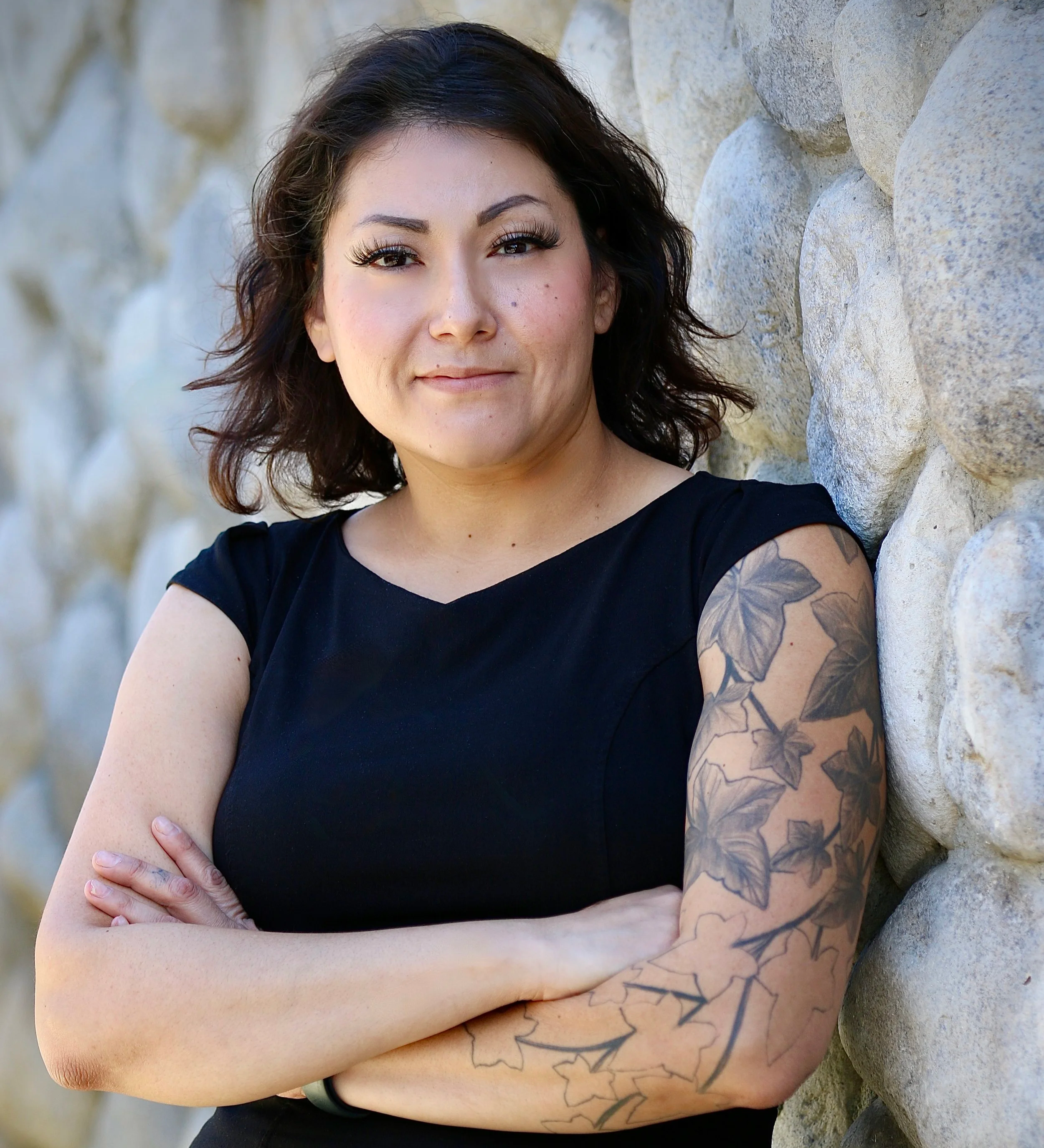 A woman with short dark hair wearing a black turtleneck and a name tag, standing outdoors near a tree, with a street and some buildings in the background.