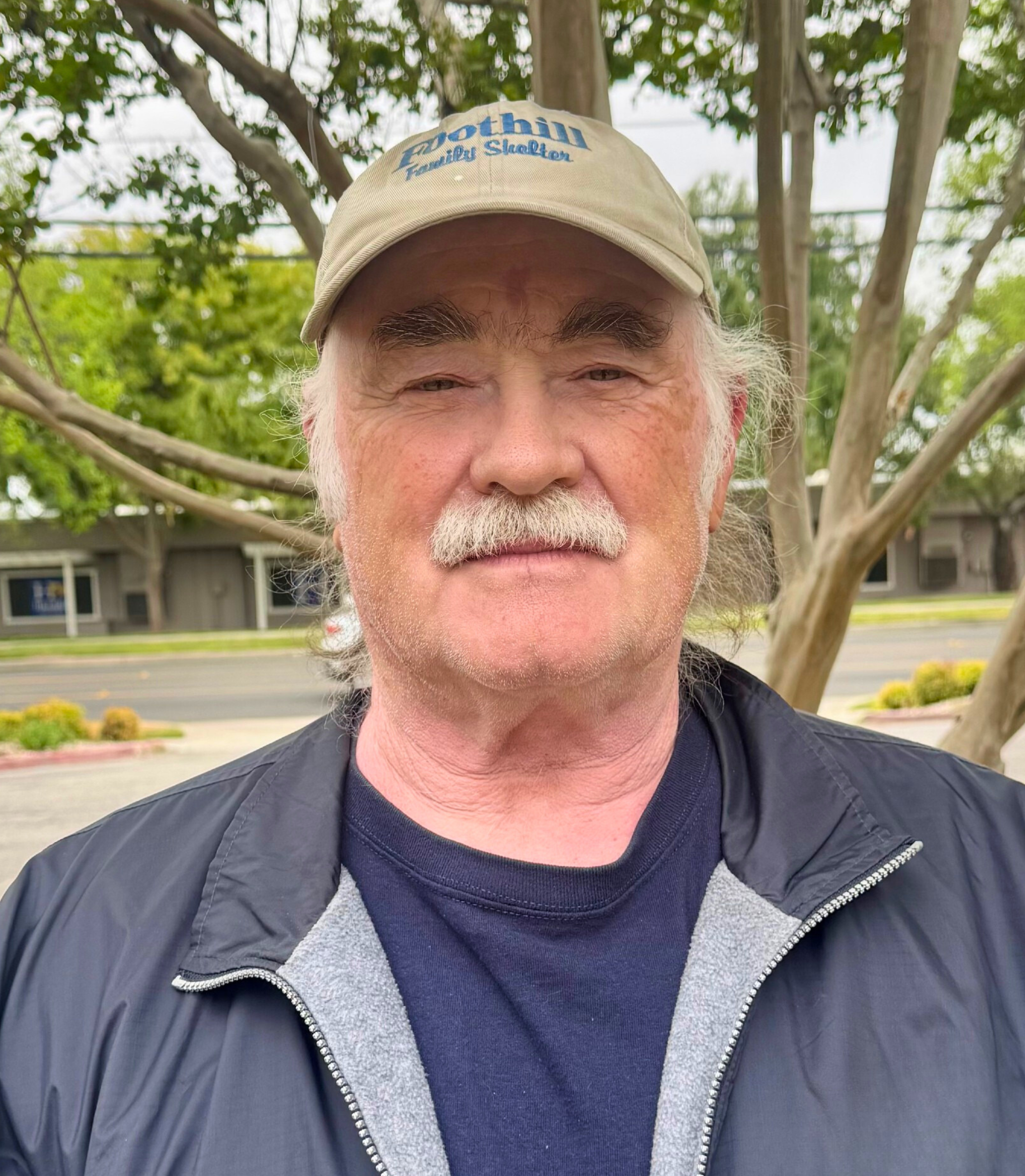 A man with gray hair and a mustache wearing a beige cap and a black jacket standing outdoors in front of trees with green leaves and a building in the background.