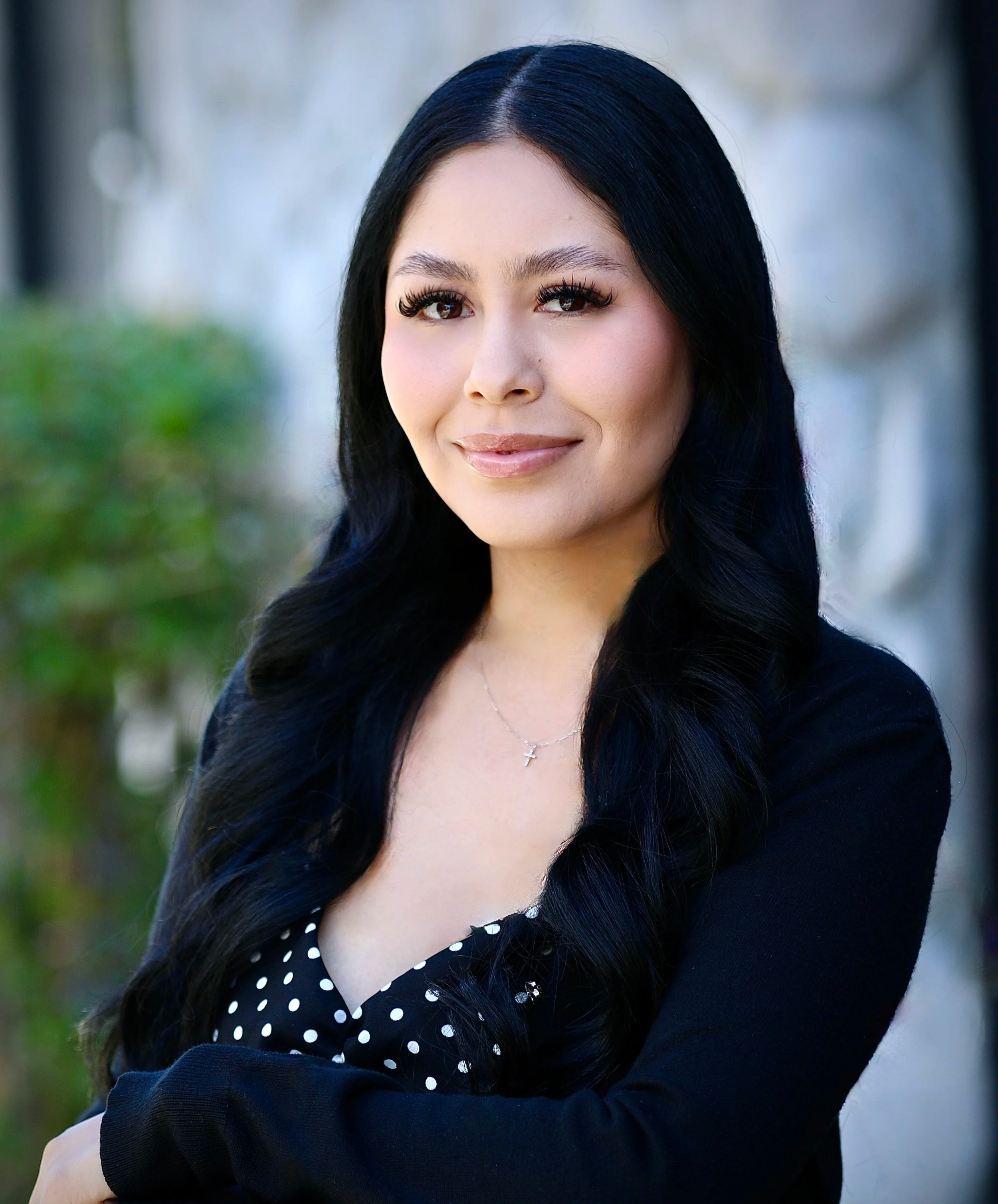A woman with long black hair wearing a black sleeveless top and a name badge standing outdoors in front of a tree with green foliage and buildings in the background.