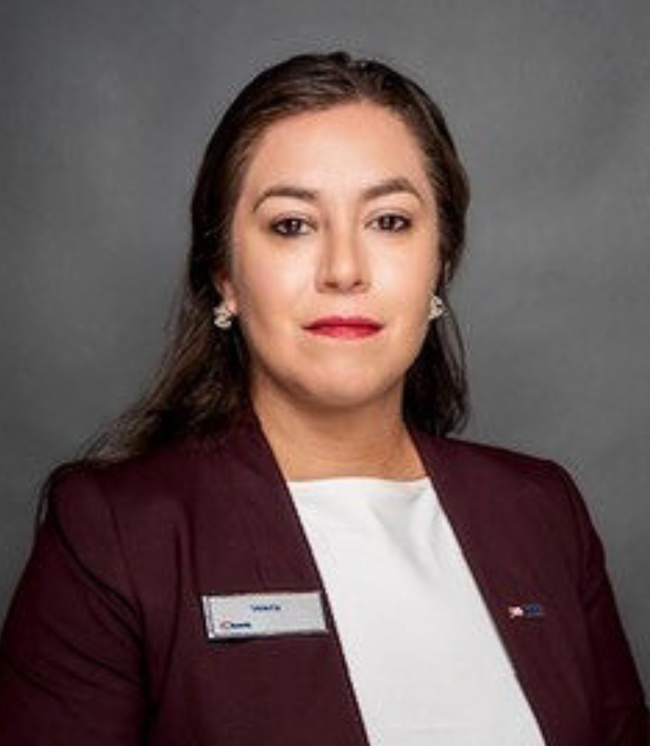 Professional woman with dark hair, wearing a maroon blazer and white blouse, posing against a gray backdrop.
