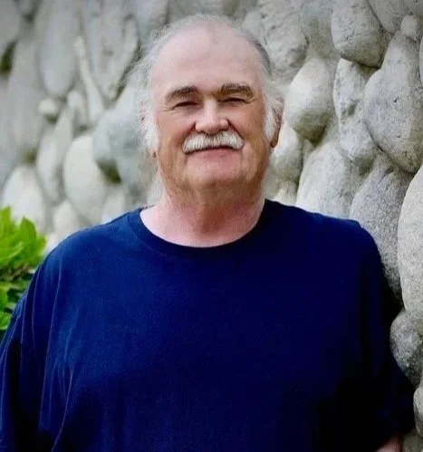 A man with gray hair and a mustache wearing a beige cap and a black jacket standing outdoors in front of trees with green leaves and a building in the background.