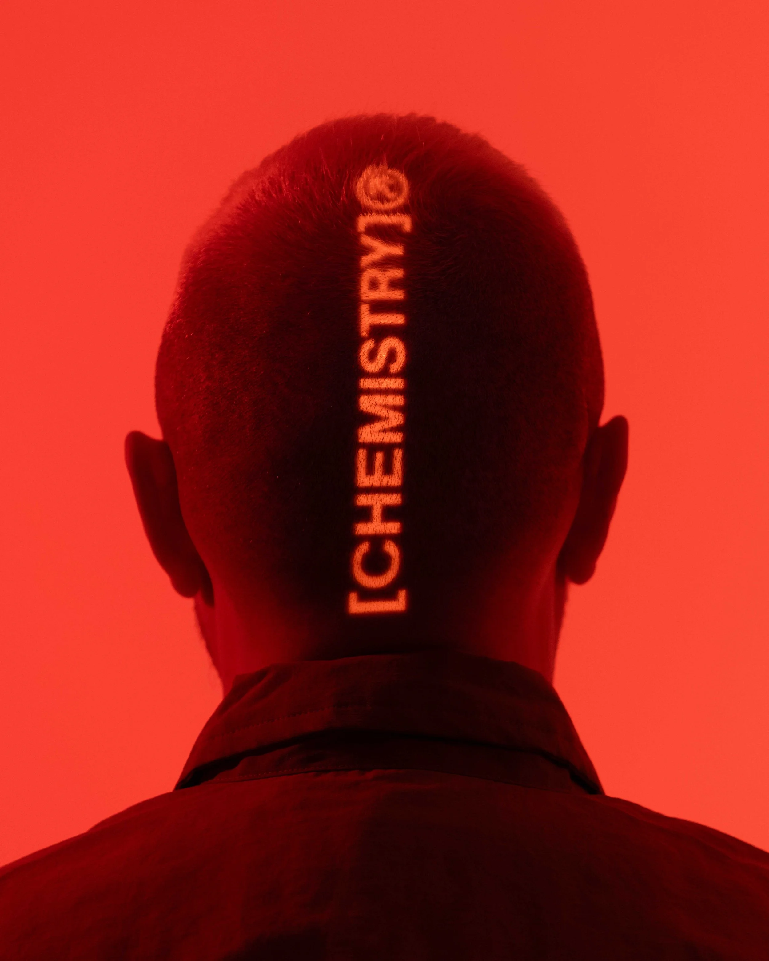 Back of a person's head with the words "LCHENMISTRT" projected vertically on their scalp in glowing orange letters, against a red-orange background.