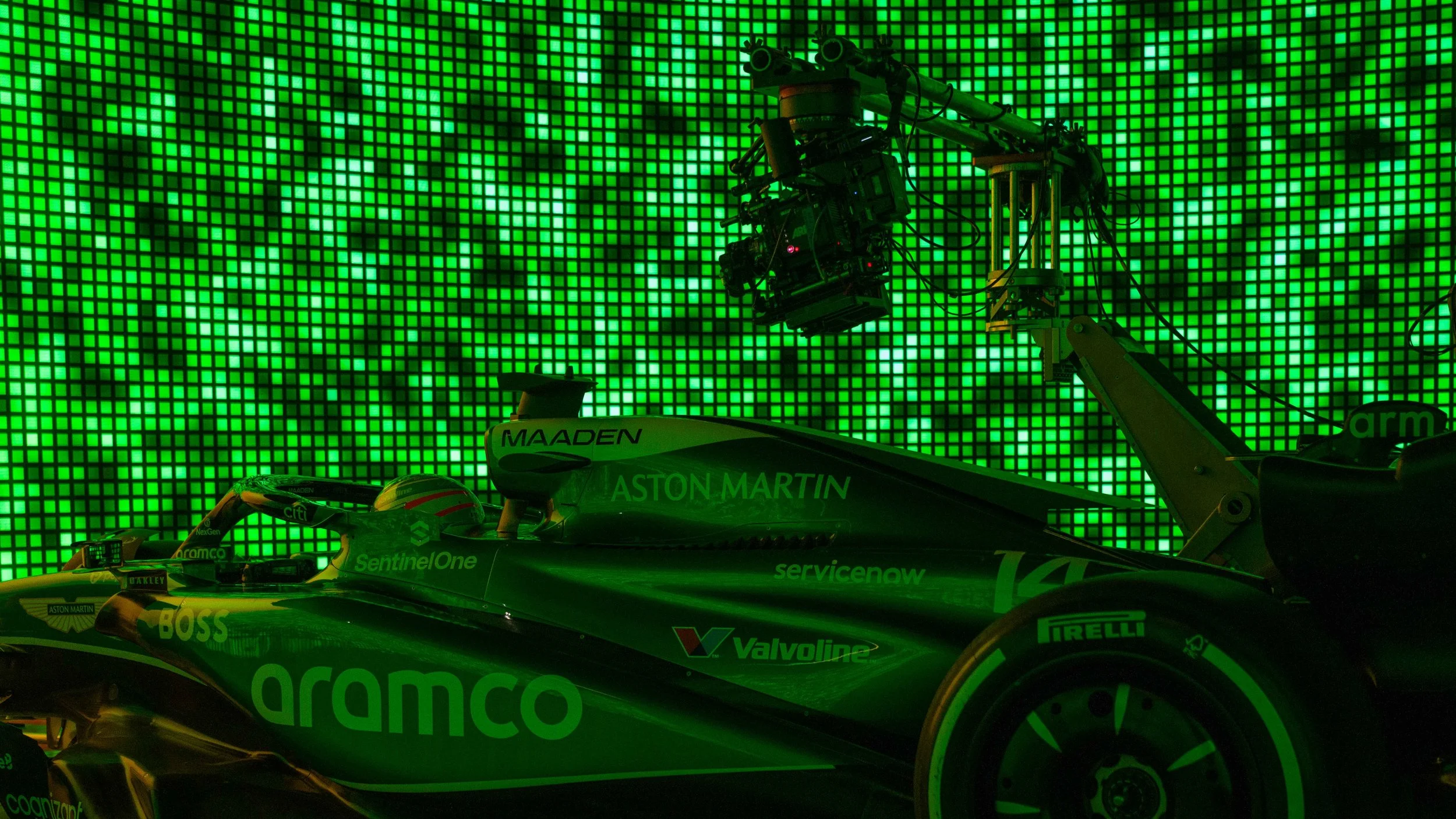 Formula 1 race car with Aston Martin branding in front of a green LED light wall. The car has sponsorship logos including Aramco, SentinelOne, Valvoline, Pirelli, and others. A camera mounted on a crane is positioned above the car, capturing footage.