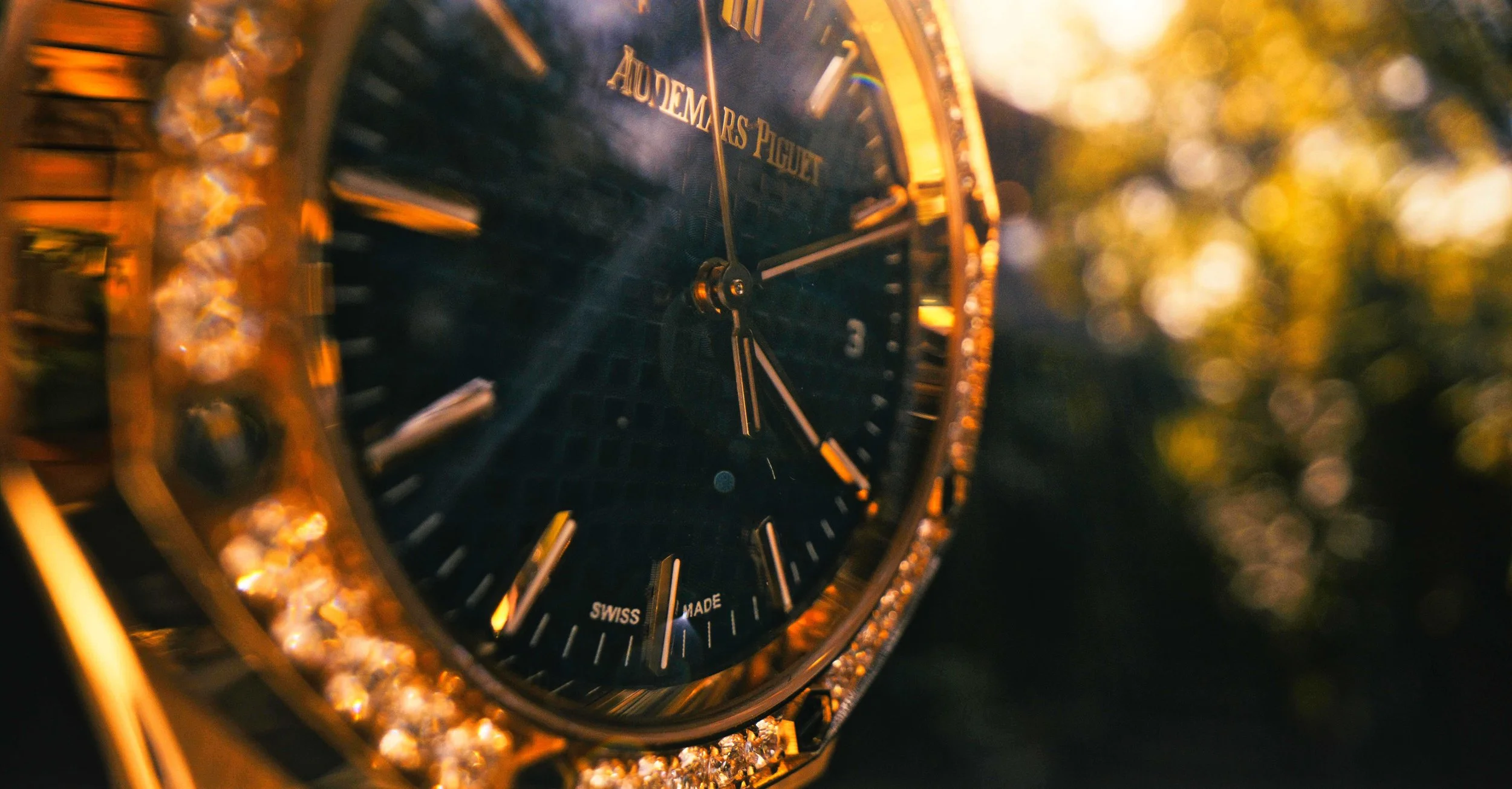 Close-up of a wristwatch with a black face, gold accents, and a border encrusted with diamonds, with trees and sunset or sunrise in the background.